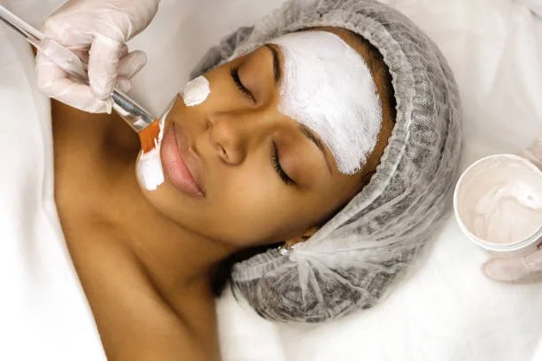 Woman receiving a facial treatment with a brush, lying down with a head cap, closed eyes, and a white facial mask on her face.