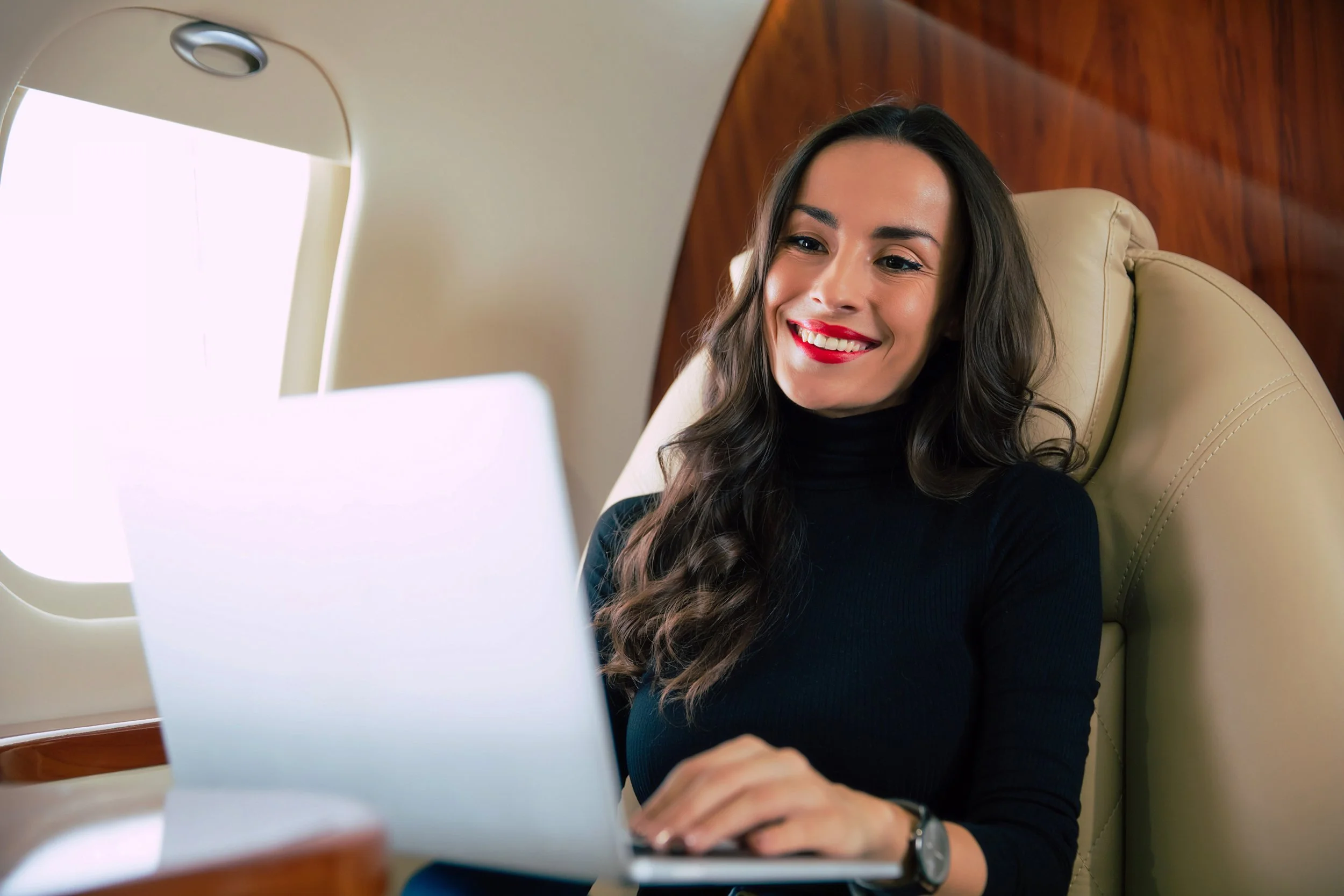 A refined woman with long dark hair and a black turtleneck shirt smiling while looking at her laptop onboard an airplane.