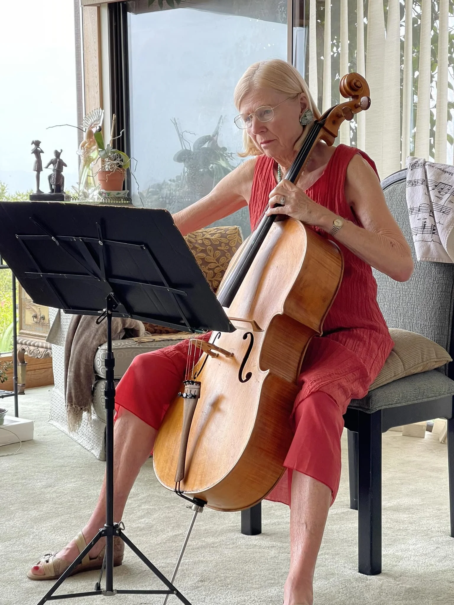An older woman with blonde hair, glasses, and statement earrings, wearing a sleeveless red top and red pants, is playing a cello indoors. She is seated on a bench with a music stand in front of her, in a bright room with large windows and various decorative items.