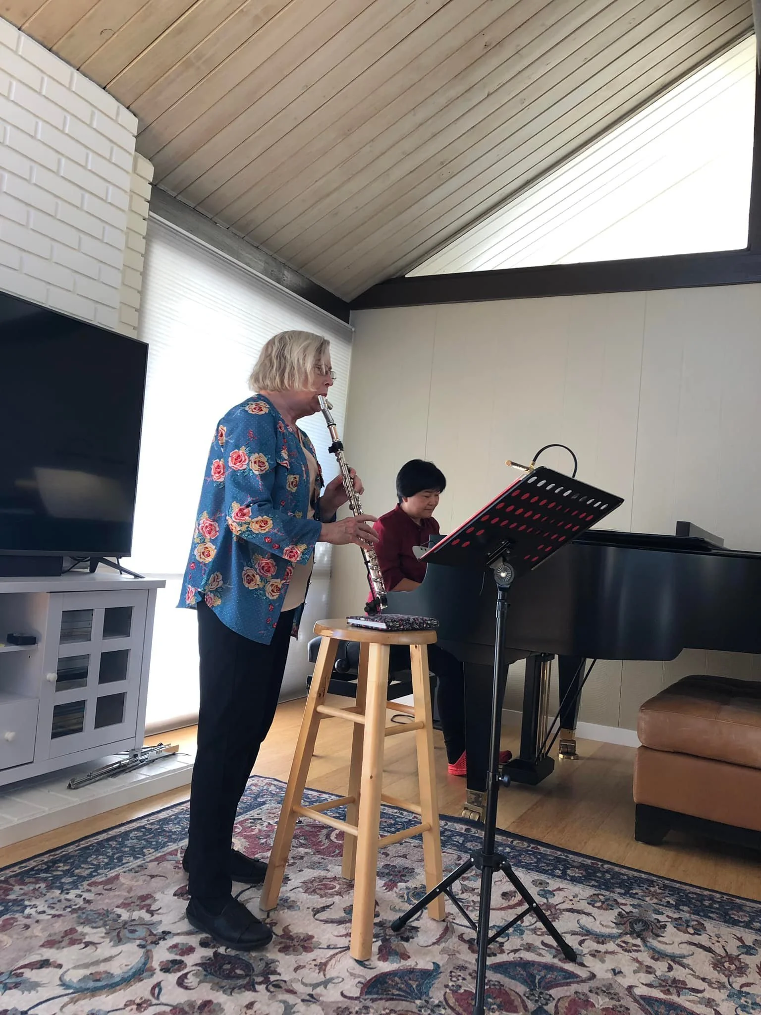 A woman playing the clarinet and a girl playing the piano in a living room with a textured rug, a mounted TV, and large windows.