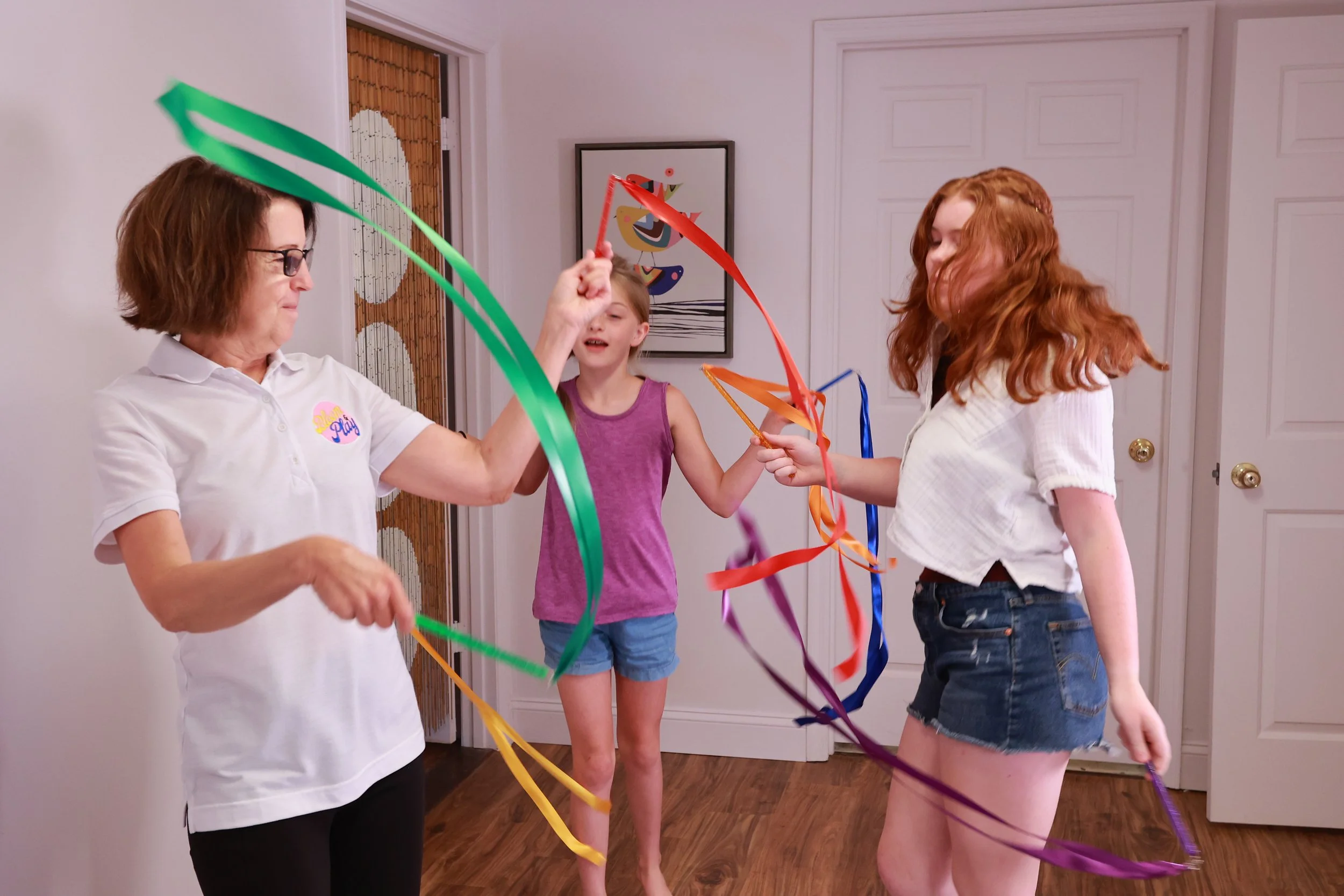 Three women and a girl dancing with colorful streamers in a room with white walls and wooden floors.