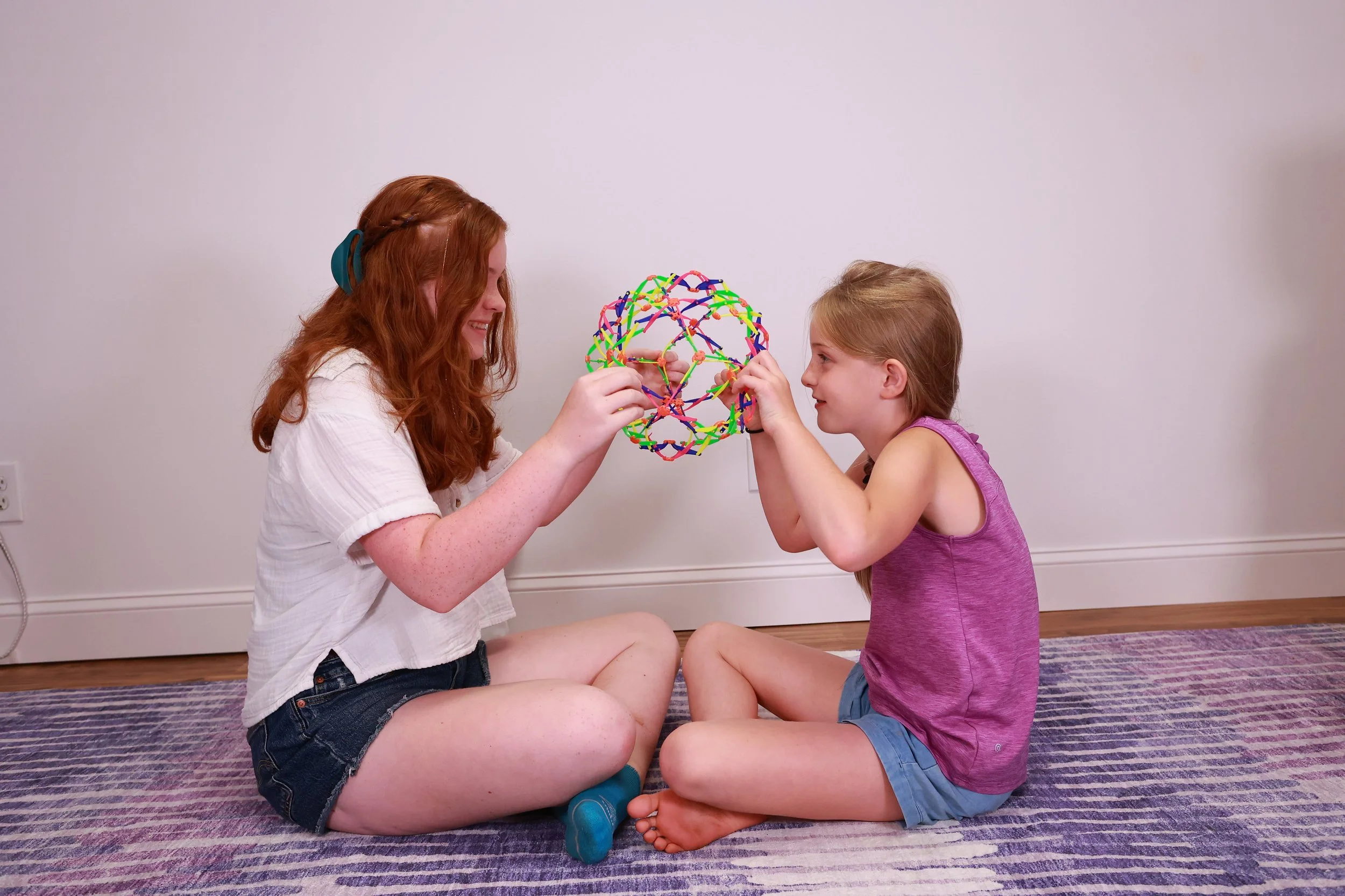 Two girls, one older with red hair and one younger with blonde hair, sitting cross-legged on a purple striped rug, play with a colorful spherical toy made of interconnected rubber bands in a plain white room.
