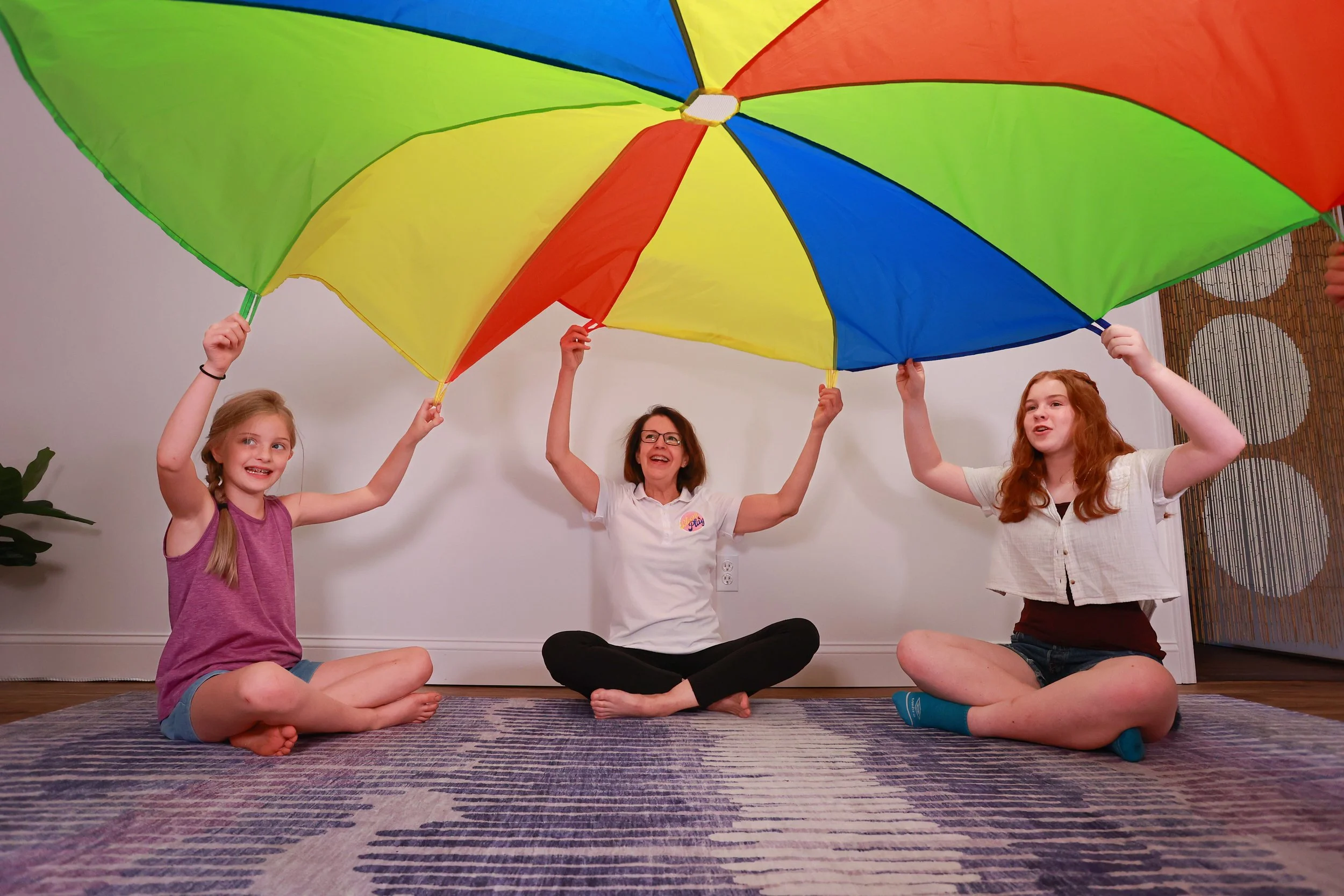 A woman and two young girls sitting on a patterned rug inside a room, holding a large colorful rainbow umbrella. The woman is in the middle, wearing glasses and a white shirt, with her arms raised holding the umbrella handle. The girl on the left has blonde hair in pigtails, wearing a purple tank top and shorts, smiling. The girl on the right has curly red hair, wearing a white shirt and shorts, also smiling. They appear to be enjoying a playful activity together.