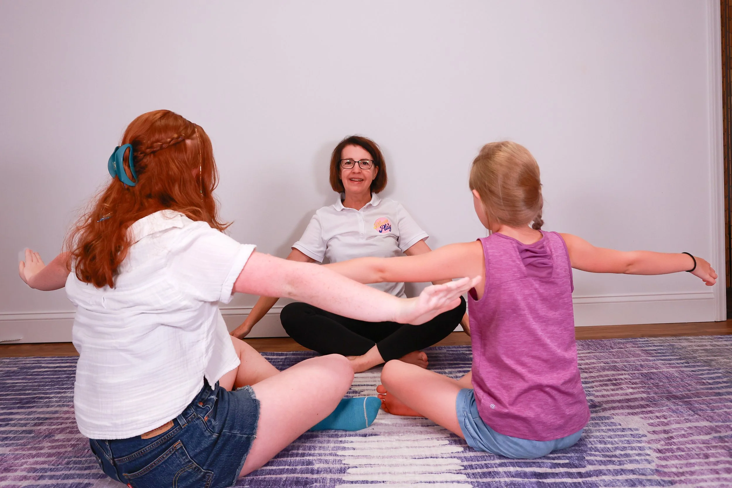 Three girls and one woman sitting on a rug, participating in a yoga or stretching session indoors. The woman, sitting in the center, is smiling and observing the girls.