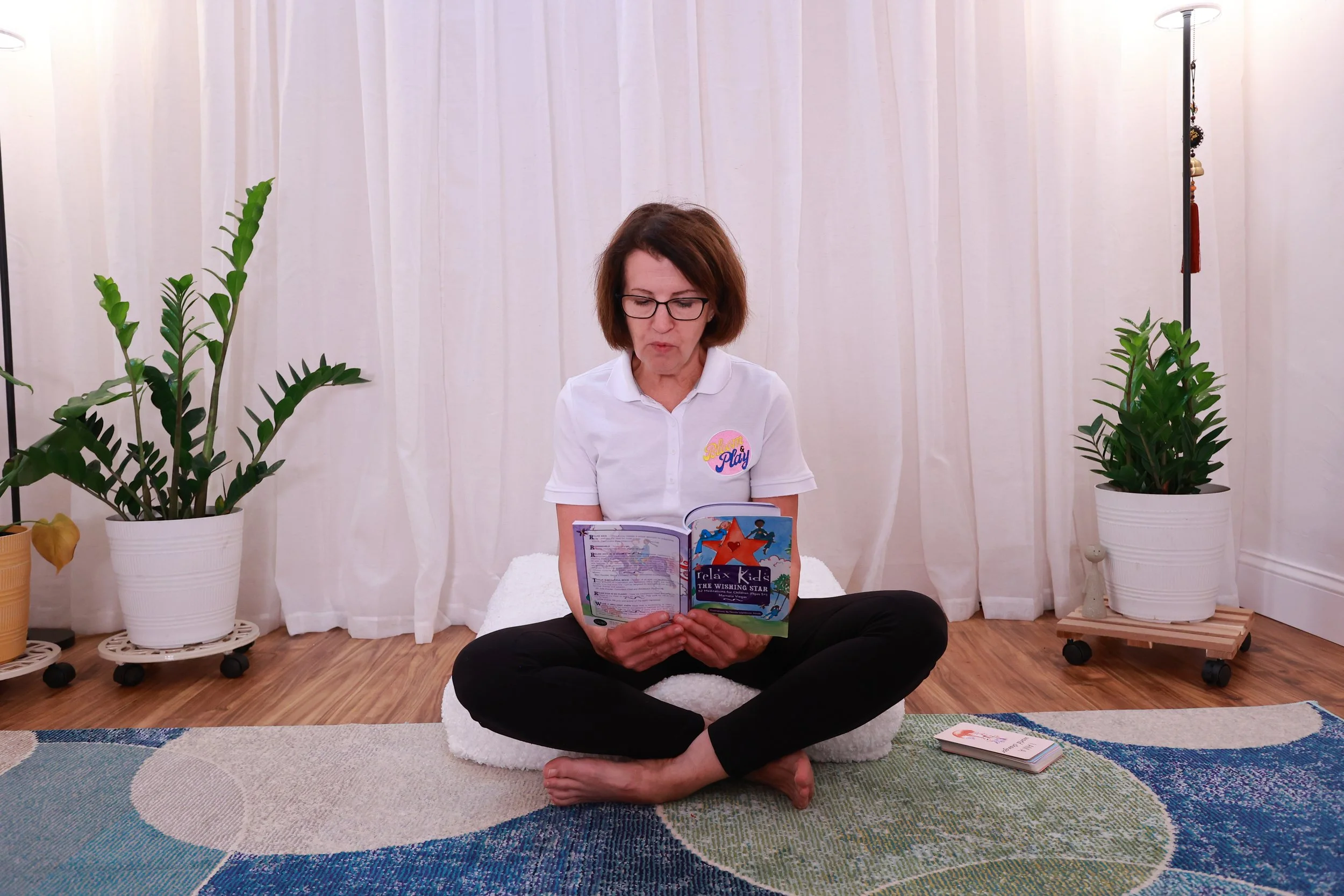 Woman sitting cross-legged on a carpet, reading a children's book in a room with white curtains, two potted plants, and a small book on the floor.