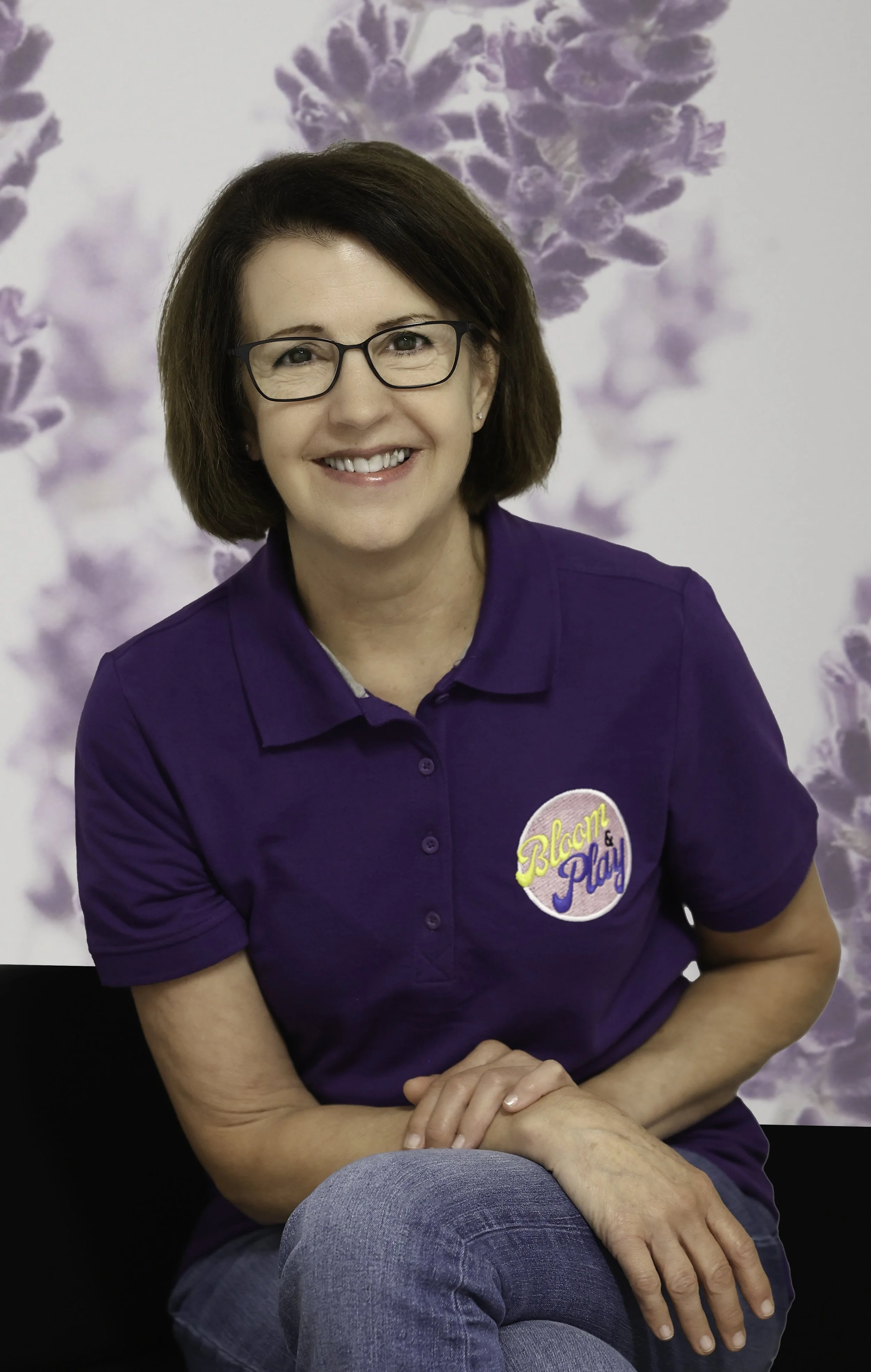 A woman with glasses and shoulder-length brown hair, smiling, wearing a purple polo shirt with a patch that says 'Blast & Play.' She is seated, with her hands resting on her lap, in front of a backdrop with purple floral designs.