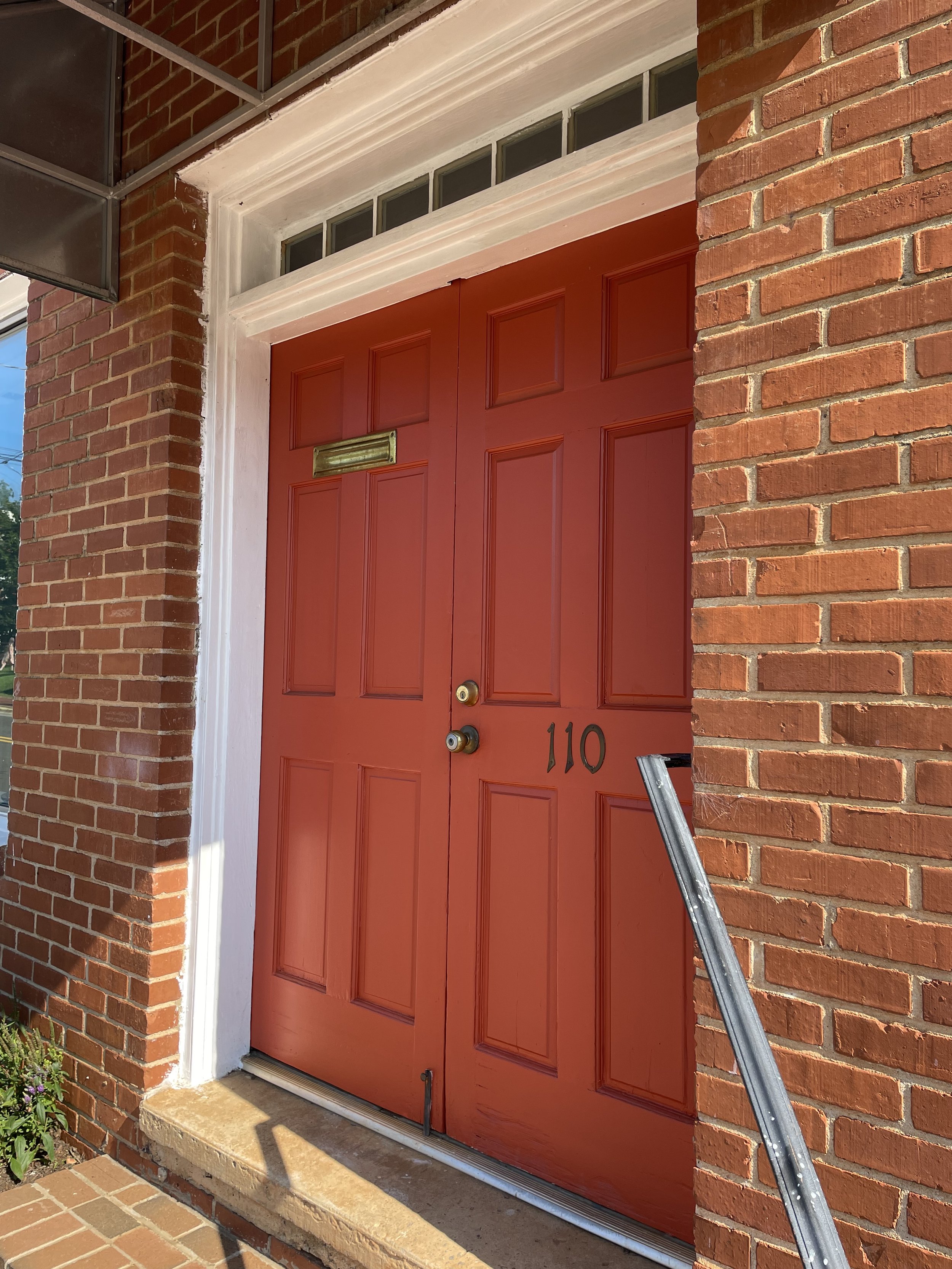 Red front door with house number 110, set in a brick wall with a white trim, a brass door handle, and a mail slot, with a metal ladder leaning against the wall on the right.