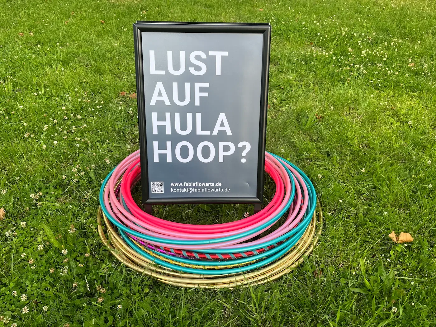 Viele Hula-Hoop-Reifen auf einer Wiese mit Werbeschild "Lust auf Hula Hoop?"