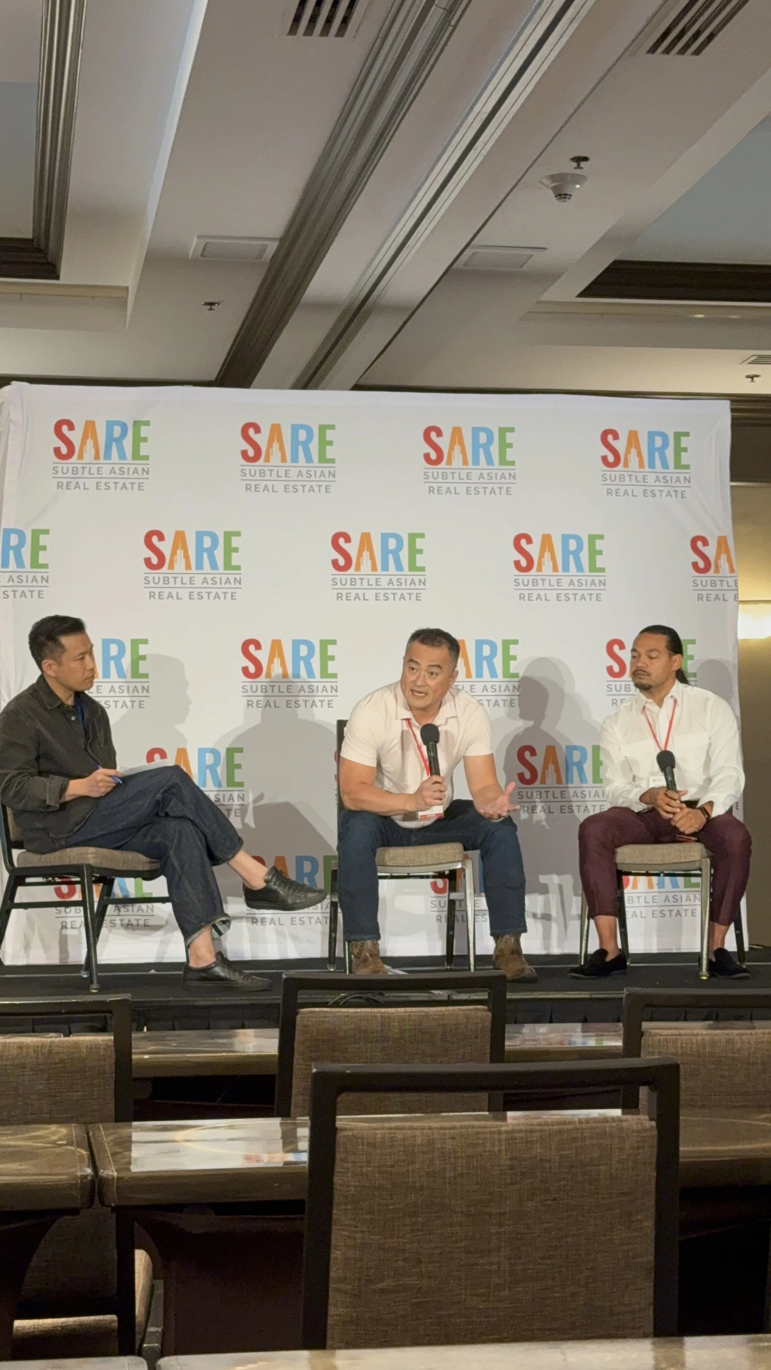 Three men sitting on a stage with a backdrop that reads 'SARE Subtle Asian Real Estate.' The man in the middle is speaking into a microphone, while the other two men listen. The stage has chairs and a black stage floor.