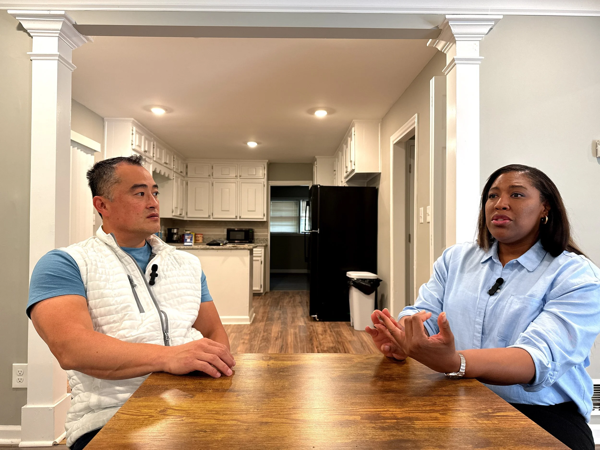 Two women sitting at a wooden table having a conversation in a kitchen with white cabinets and black appliances.