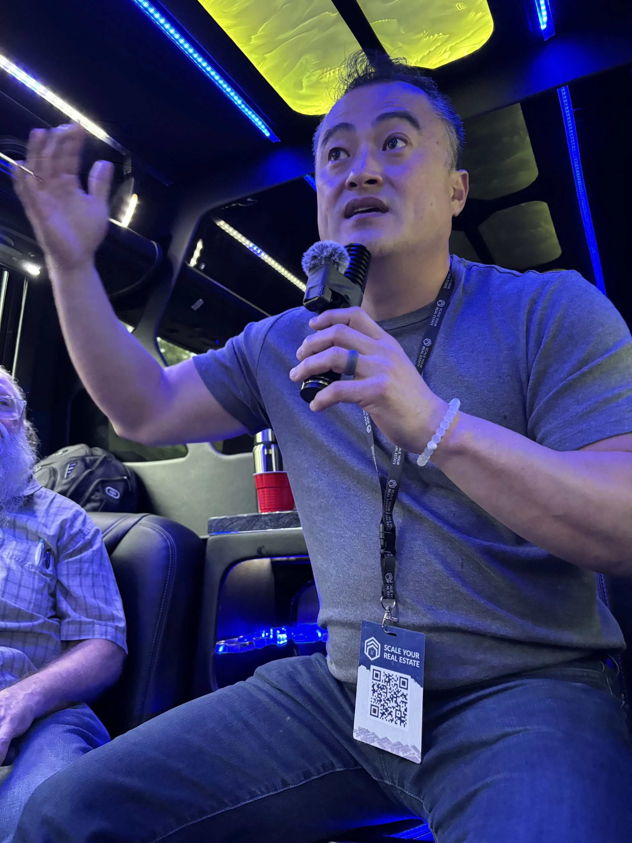Man speaking into a microphone while sitting on a bus or vehicle with blue and yellow lighting, wearing a lanyard with a QR code and the text 'Scale Your Real Estate'.