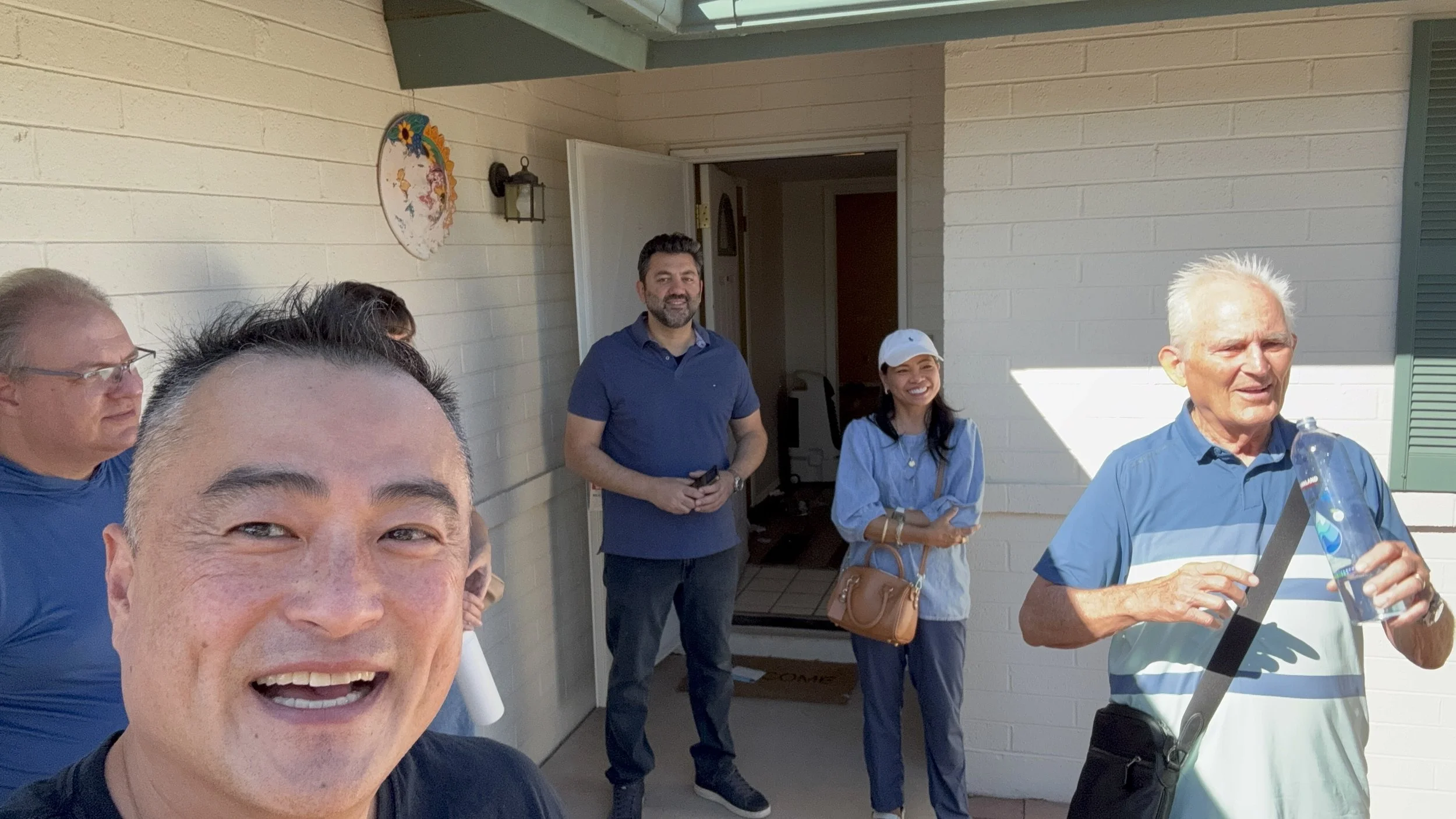 Group of five people standing on a porch outside a house with white brick walls, smiling and talking. One elderly man is holding a water bottle. The others include a woman wearing a white cap, a man in a blue shirt, and a woman with dark hair holding a brown purse. A man in front is taking a selfie.