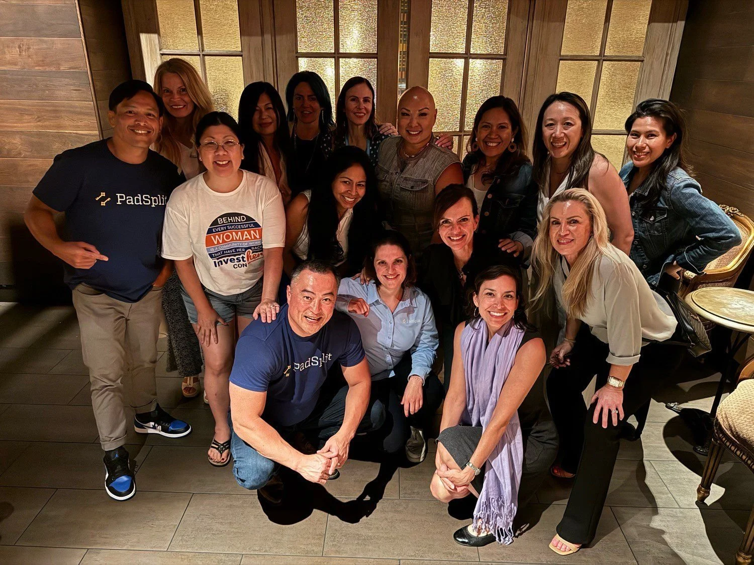 Group of 16 women and 1 man smiling and posing for a photo inside a wooden-paneled room with windows in the background.