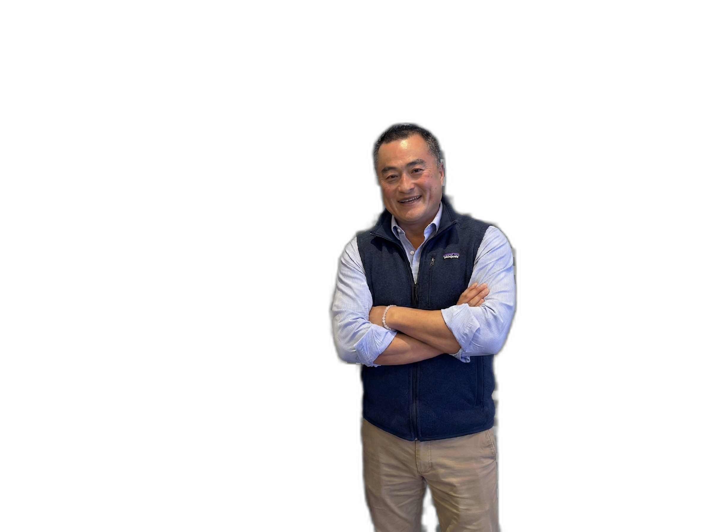A man with short black hair smiling and standing with arms crossed, wearing a light blue shirt and a dark blue vest against a black background.