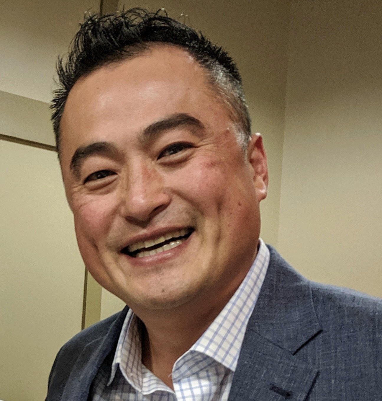 A smiling man in a gray suit and checkered shirt standing in an indoor setting.