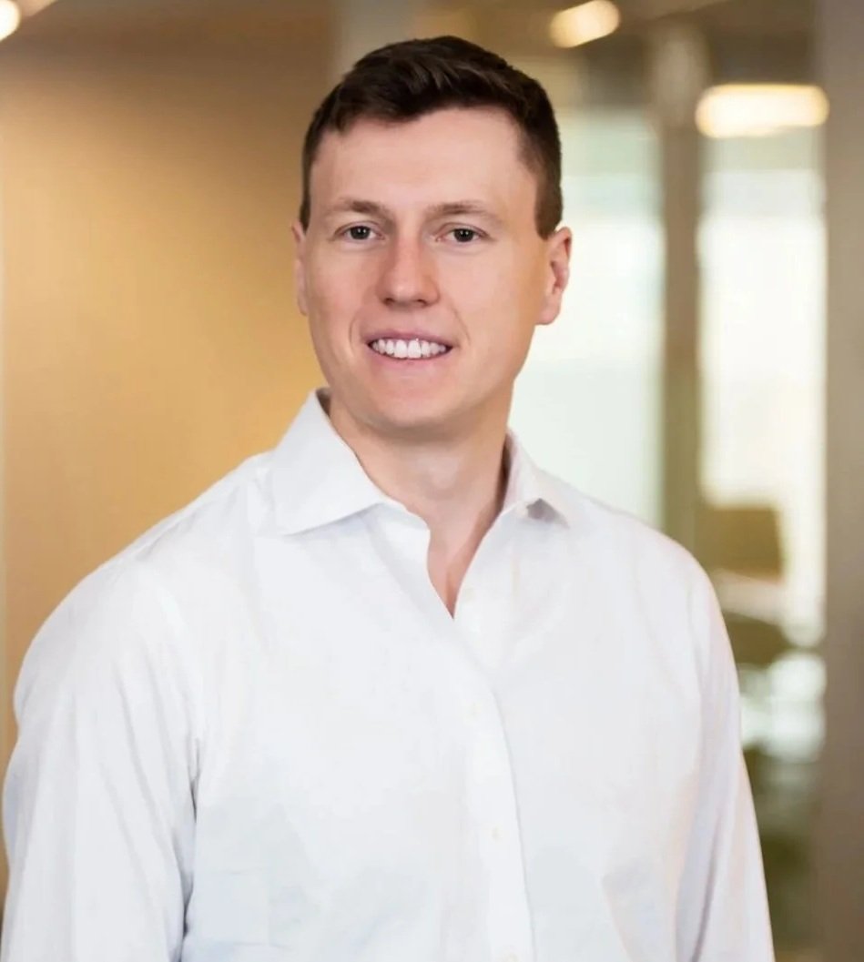 A young man with short brown hair smiling, wearing a white shirt, standing in an indoor setting with a blurred background of windows and lights.