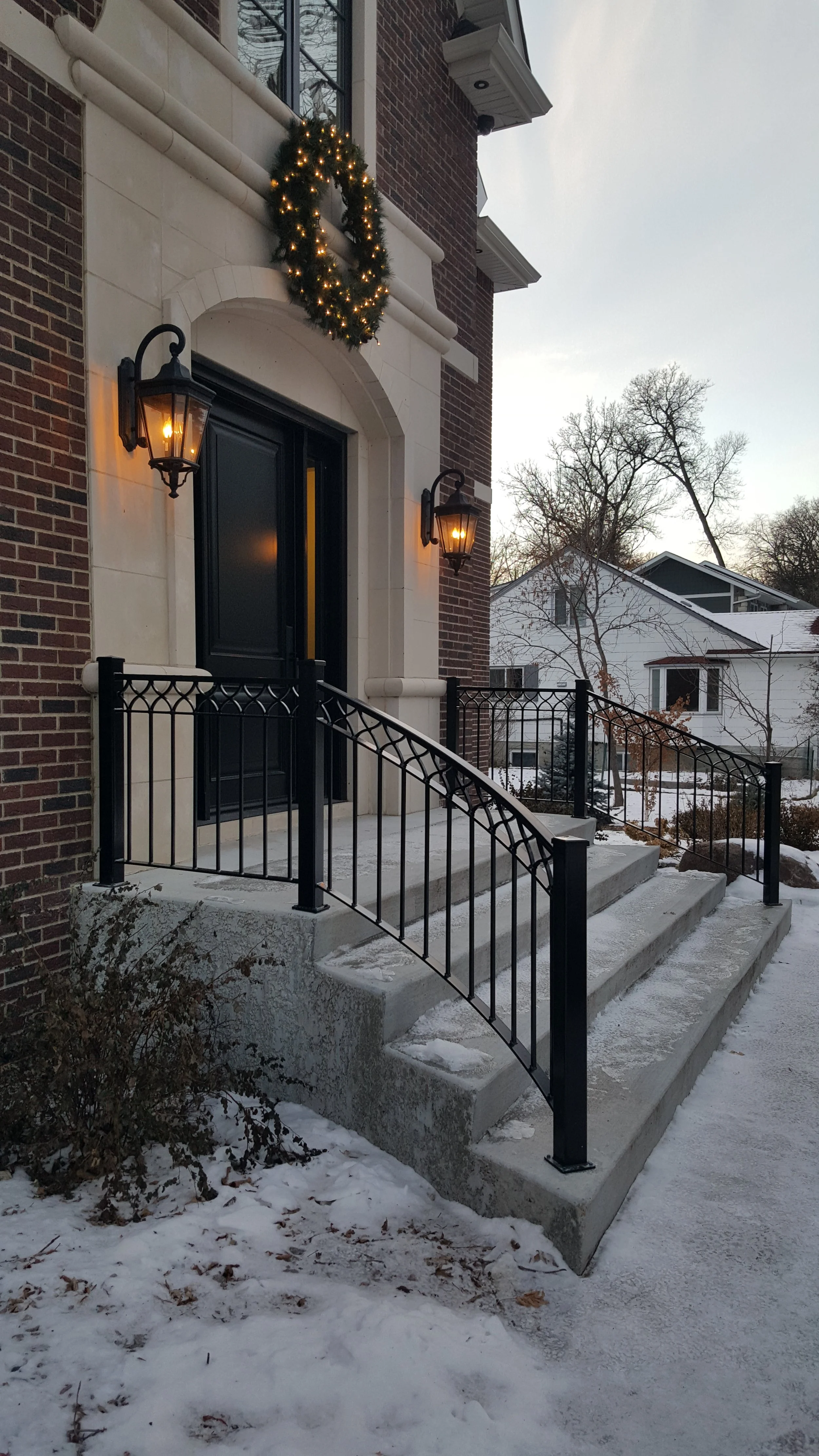 Front porch of a house with concrete stairs, black metal railing, black double doors, and two wall-mounted lanterns lit at dusk. A holiday wreath with lights is above the door. Snow covers the ground and surrounding landscaping.