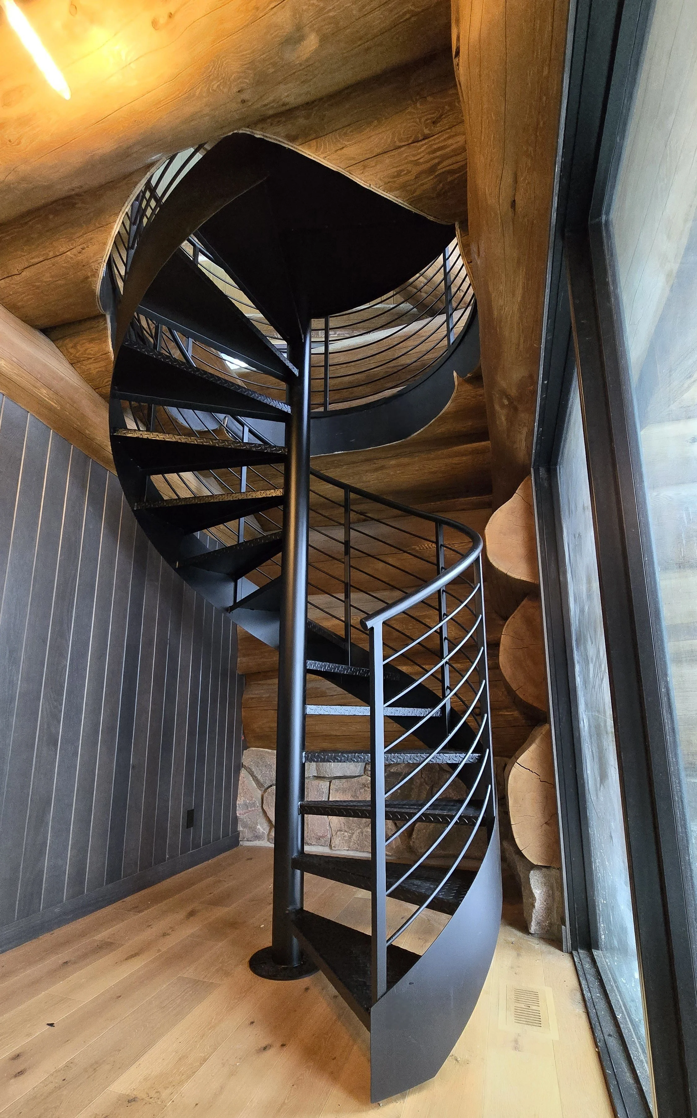 Interior view of a black metal spiral staircase with wooden walls and flooring, next to a large glass window.