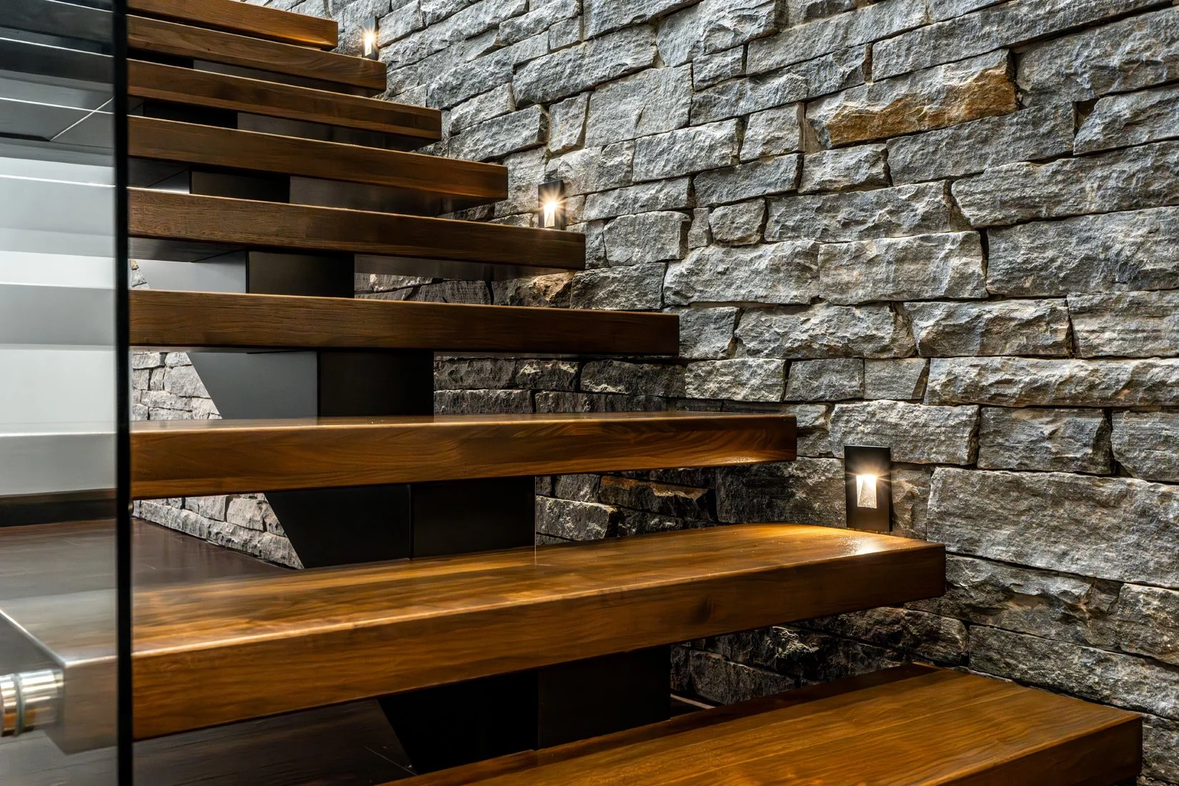 Close-up of a modern staircase with dark wood steps, metal supports, and a textured stone wall background.