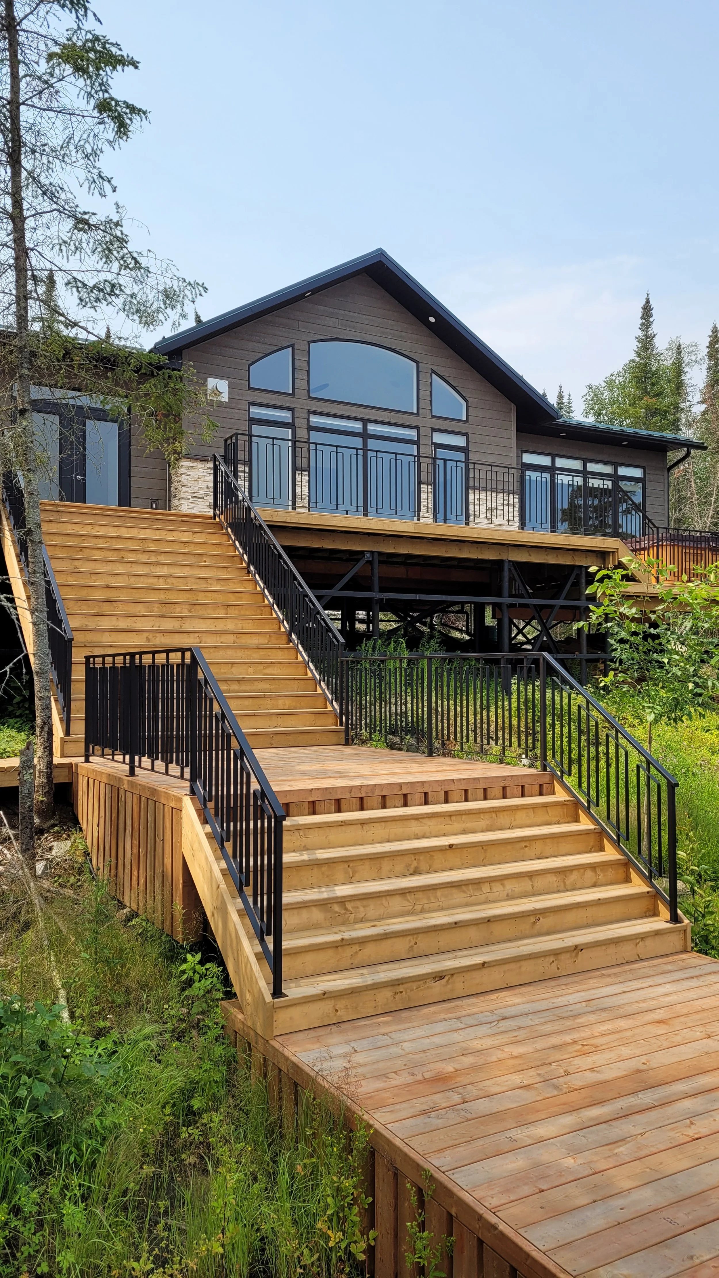 A modern house with a large deck and stairs leading up to it, surrounded by greenery.