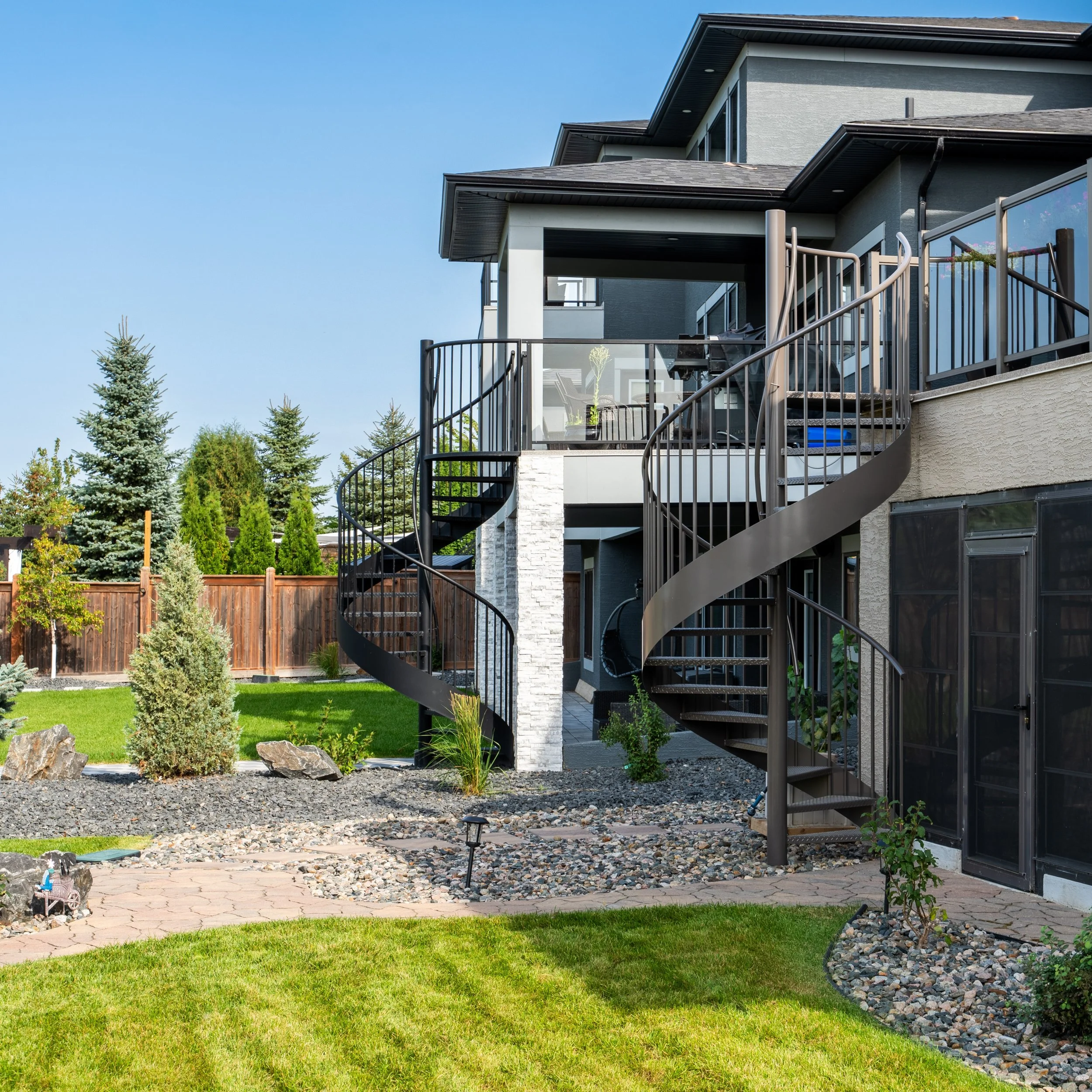 Modern multi-story house with backyard featuring a spiral staircase, green lawn, and landscaped garden with rocks, small trees, and shrubs.