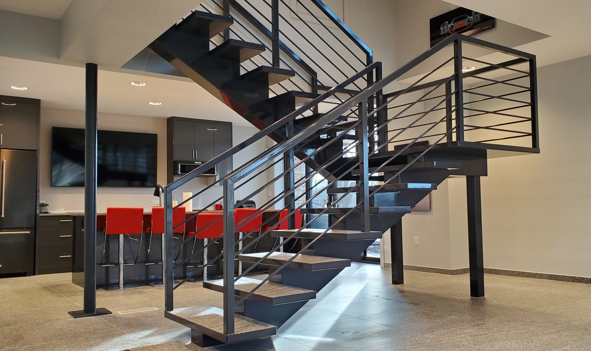 Interior view of a modern staircase with black metal railing and wooden steps in a contemporary home. The background features a kitchen with dark cabinets, red barstools, and a large flat-screen TV mounted on the wall.