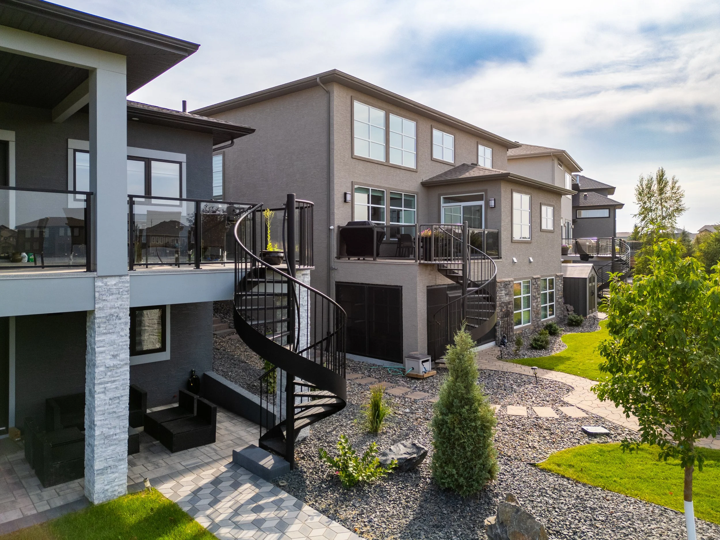 Modern multi-story residential buildings with outdoor decks, spiral staircases, a landscaped yard with trees, grass, and a walkway, under a partly cloudy sky.