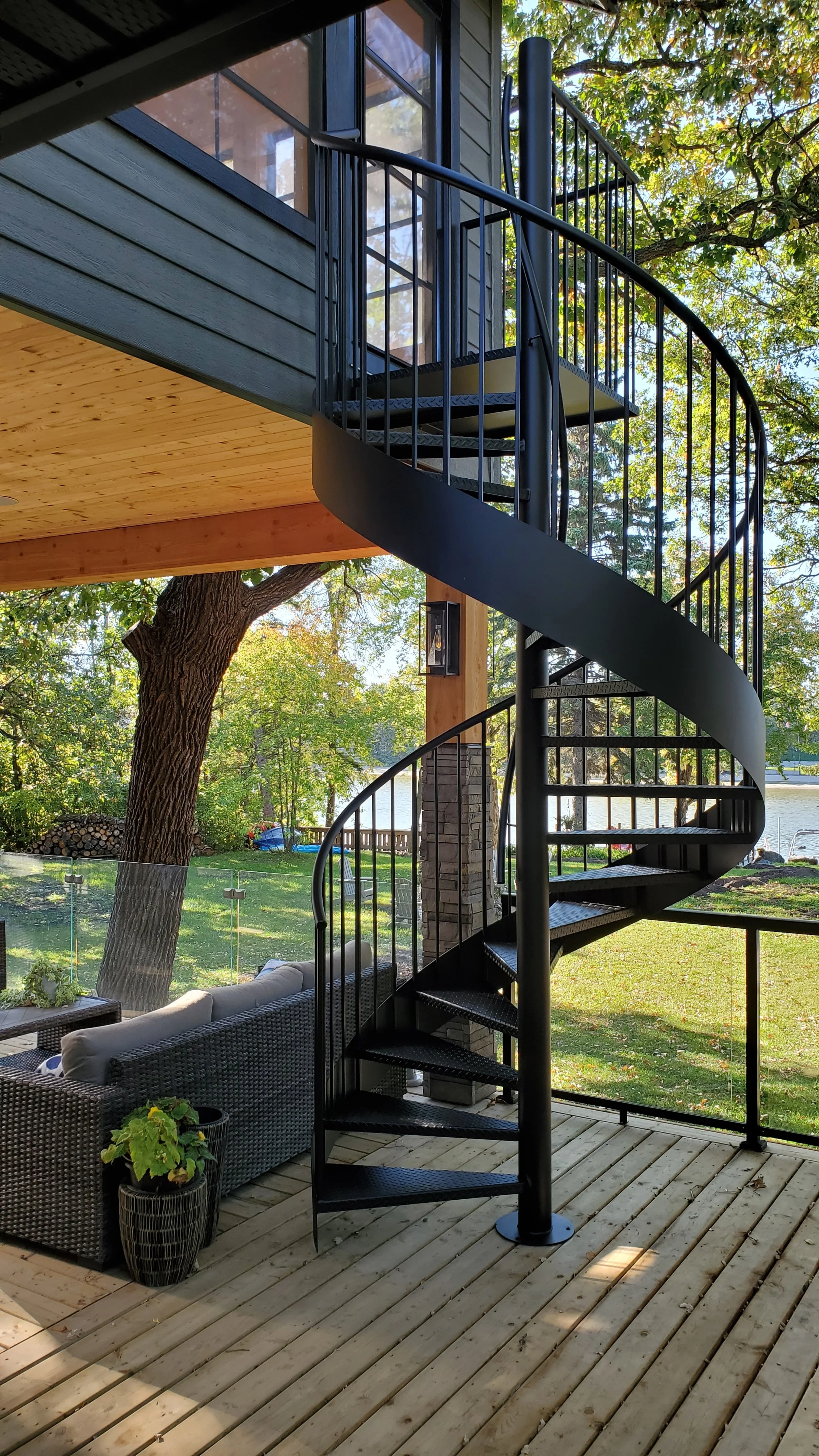 A black spiral staircase on a deck outside a house, with a view of trees, a lake, and outdoor furniture, during daytime.