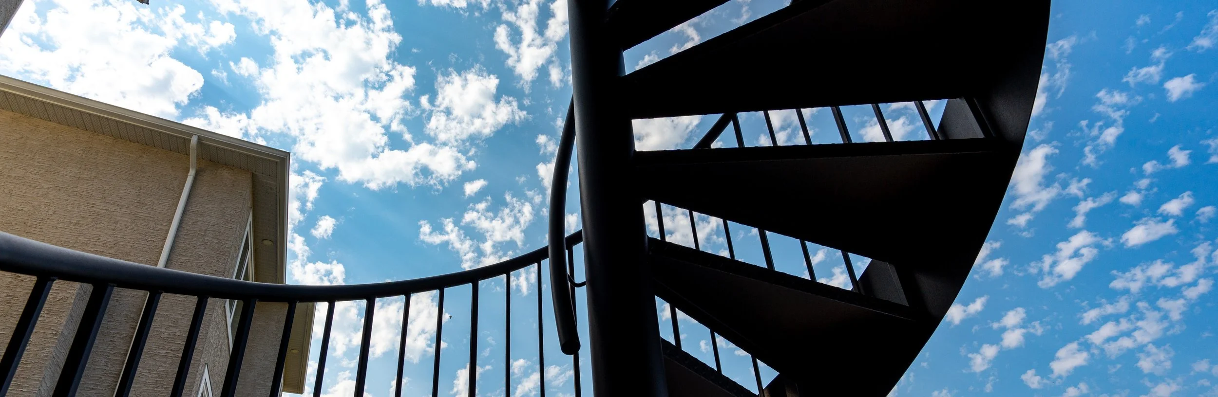Looking up at a black outdoor spiral staircase against a bright blue sky with scattered white clouds, adjacent to a beige building.
