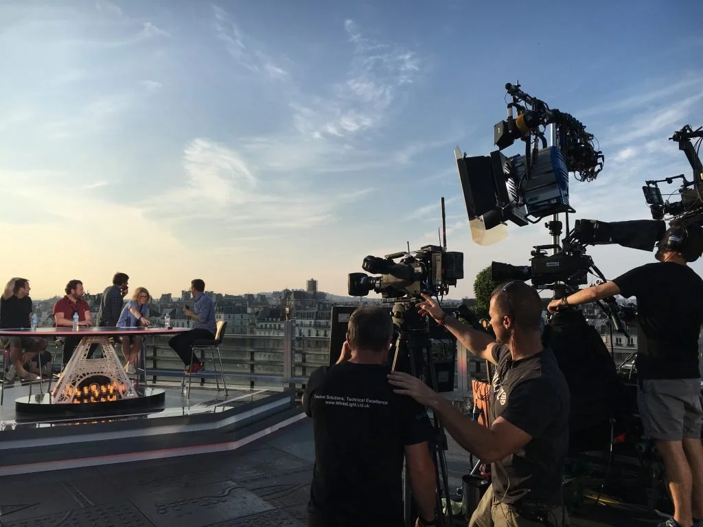People participating in an outdoor interview or talk show on a rooftop with a cityscape in the background, surrounded by camera equipment and crew members during sunset.
