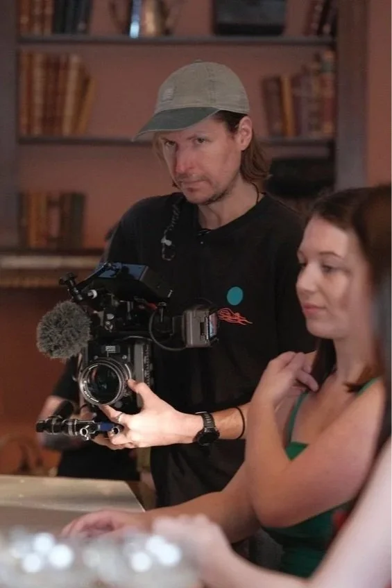 A man in a baseball cap operating a professional video camera with a woman sitting in front of him in a room with bookshelves.