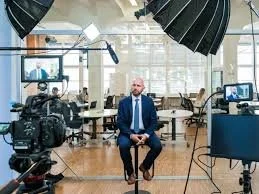 Man in a suit sitting in a modern office during a video interview or recording, with professional camera and lighting setup.