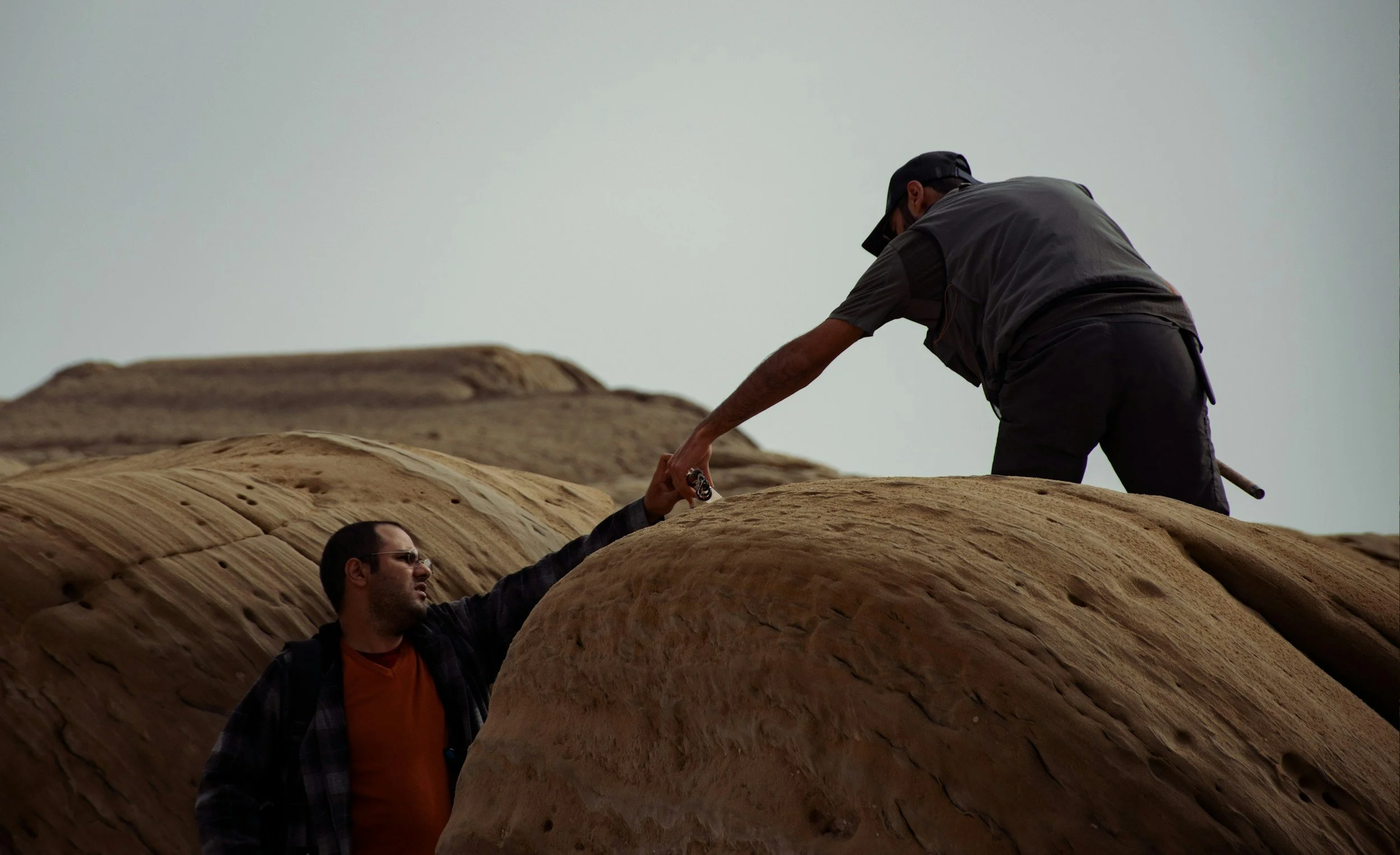 Two men climbing and helping each other on large sandstone rock formations in a desert environment.