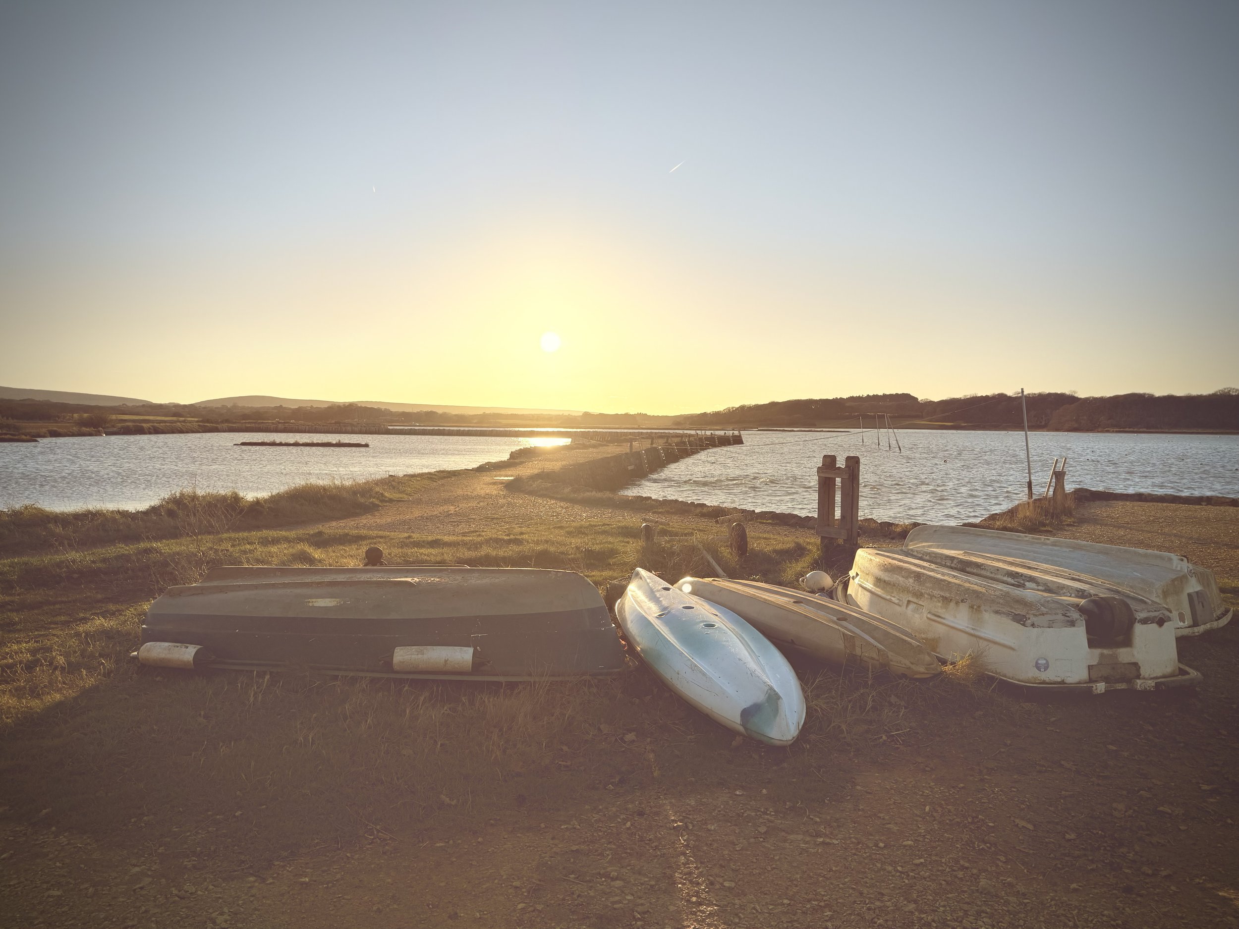 A lakeside scene at sunset with overturned boats on the ground, a waterway extending into the distance, and hills on the horizon.
