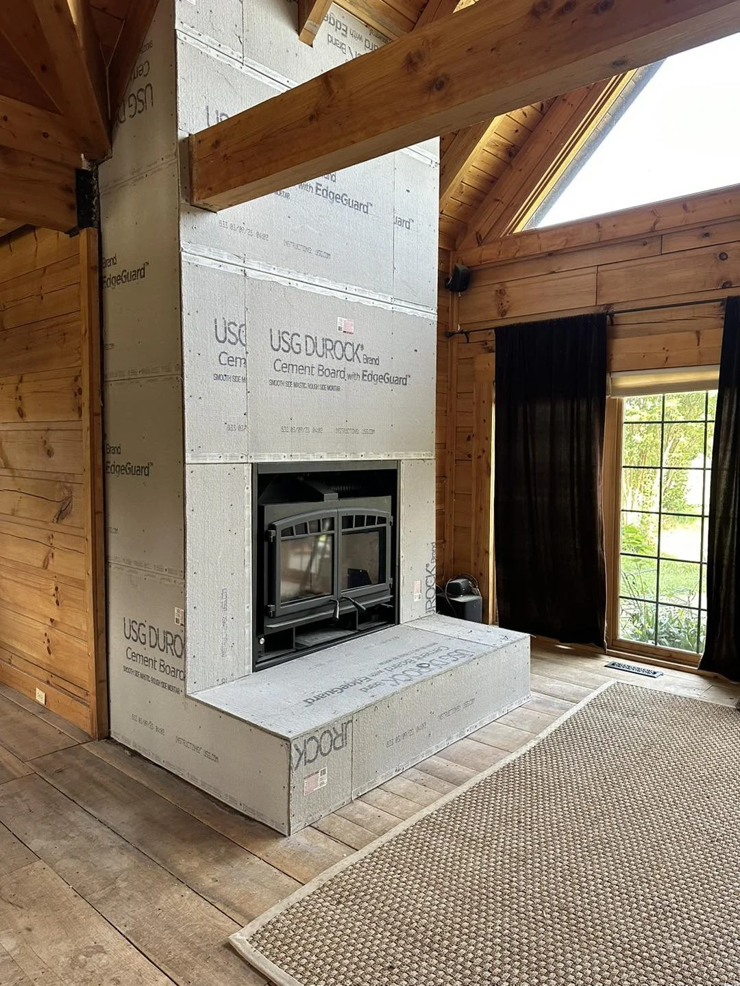 An unfinished indoor fireplace surrounded by construction insulation, with a wooden wall, ceiling, and floor, near a large window with black curtains.