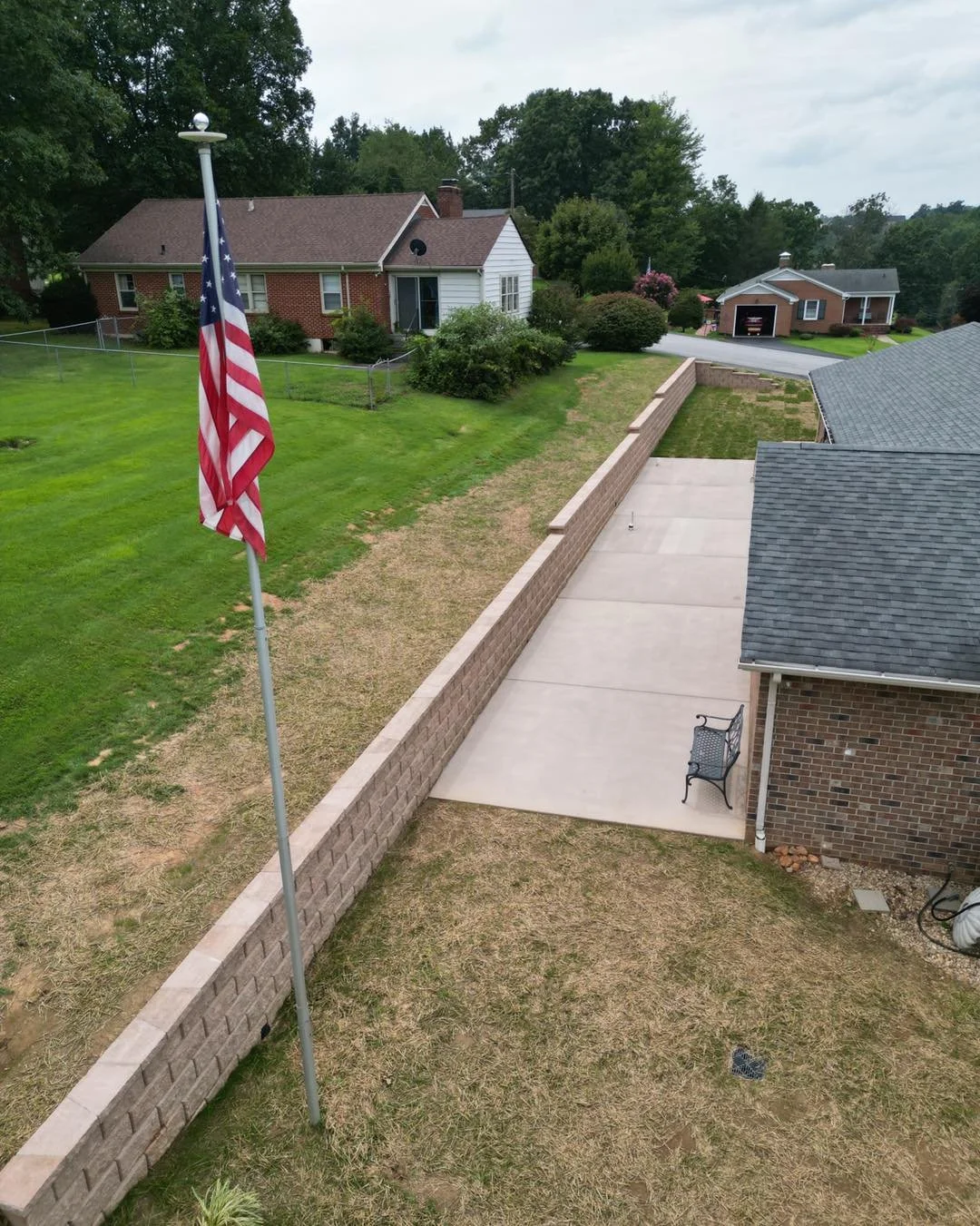 A backyard view with a flagpole and American flag, brick wall, concrete patio, grassy area, and neighboring houses surrounded by trees.