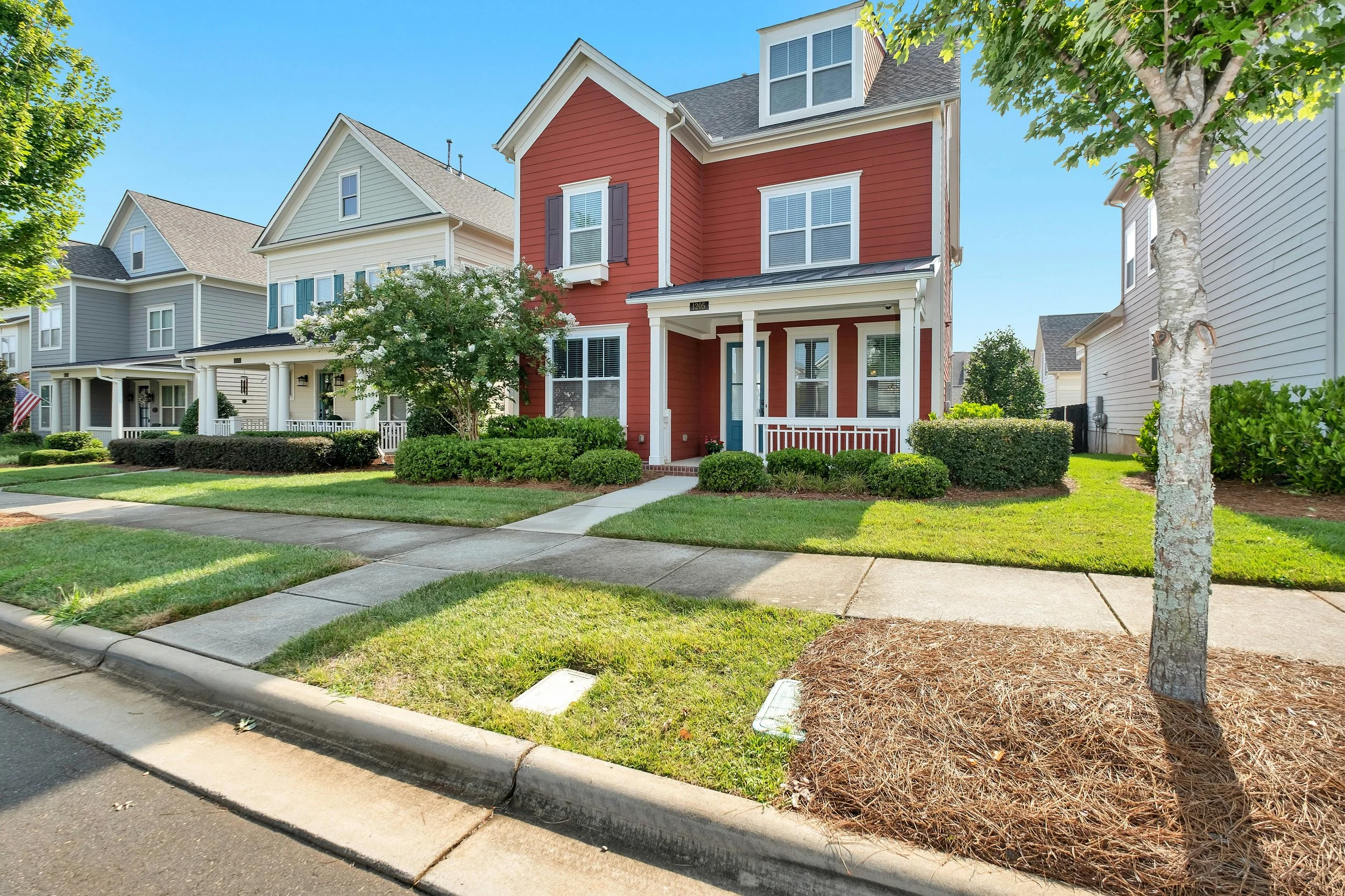 A row of colorful suburban houses with front yards, trees, and sidewalks on a sunny day.