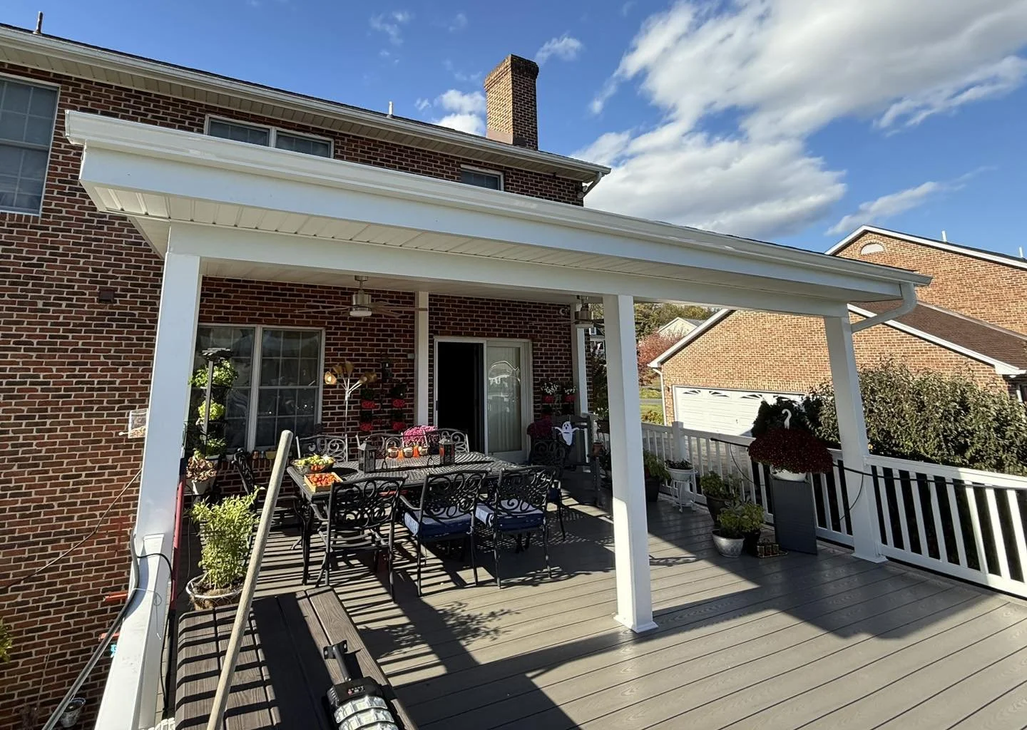 Backyard patio with metal dining table and chairs, potted plants, and hanging flower baskets on a wooden deck. Brick house with sliding glass door, gabled roof, and chimney under a partly cloudy sky.