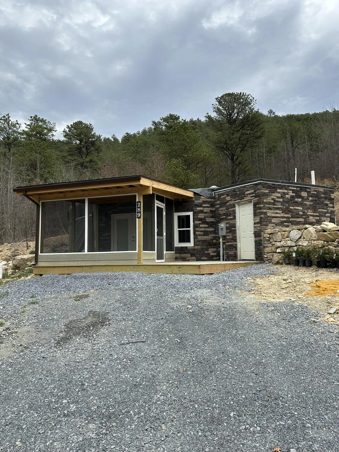 Newly constructed small house with a screened porch, dark stone exterior, white door, and surrounded by gravel driveway, with trees and cloudy sky in the background.