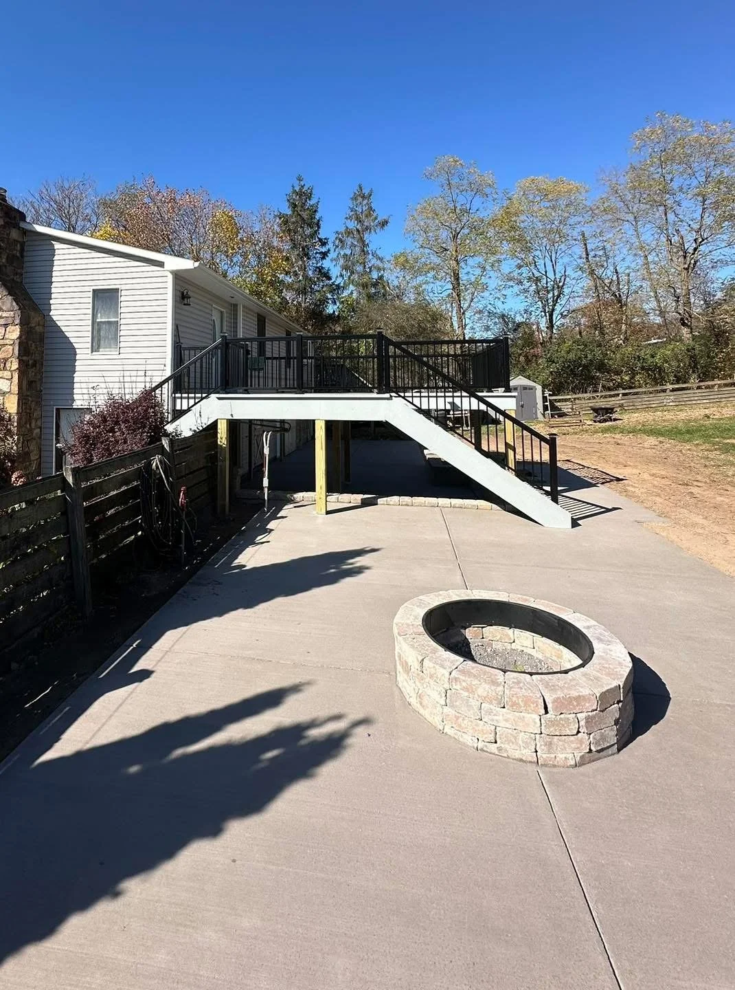 Backyard with a concrete patio, a circular fire pit made of bricks, and a elevated deck with black railing stairs leading down. House with light siding and window, trees in the background under a clear blue sky.