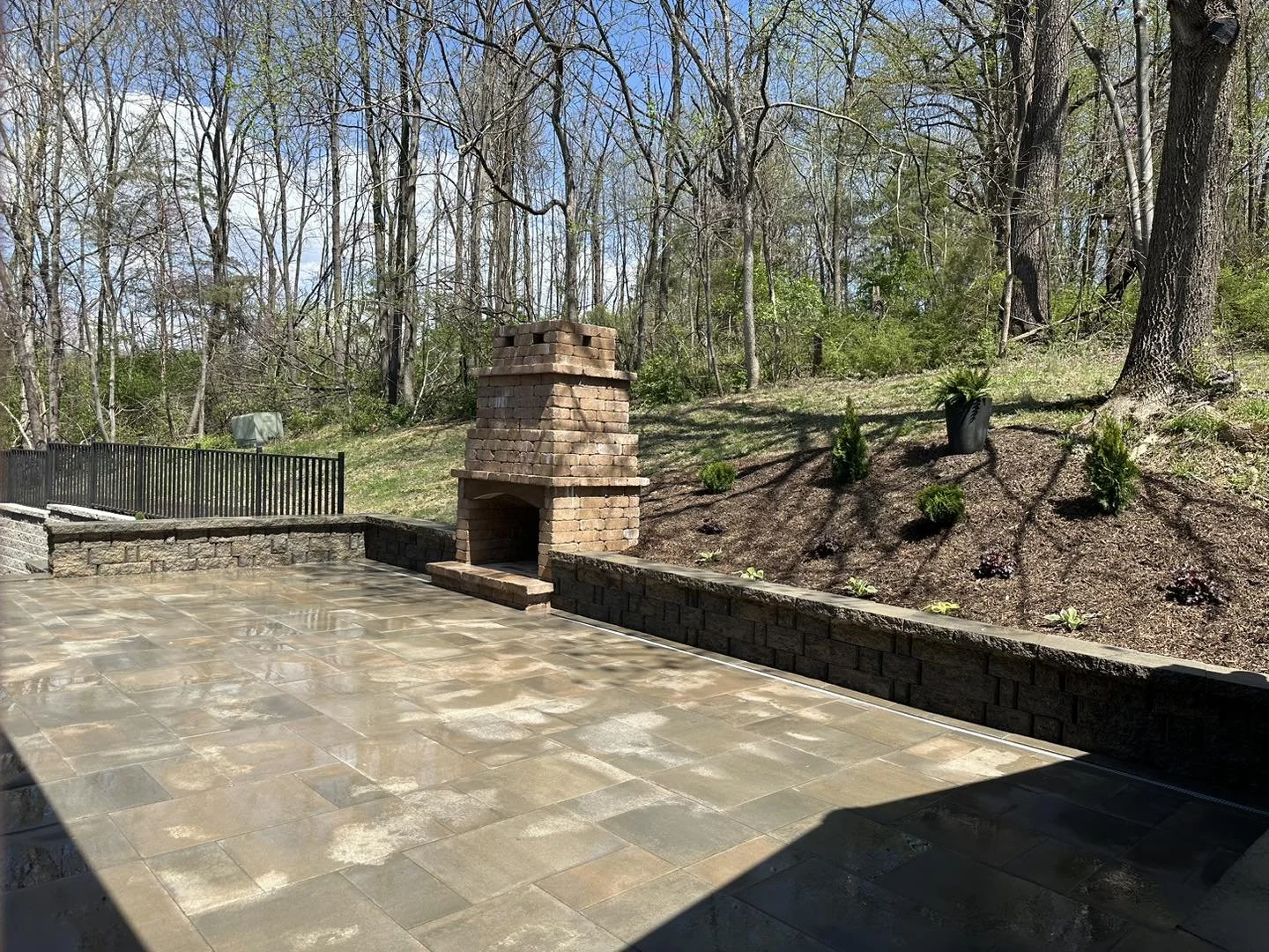 Outdoor patio with brick fireplace, stone wall, and surrounding trees.