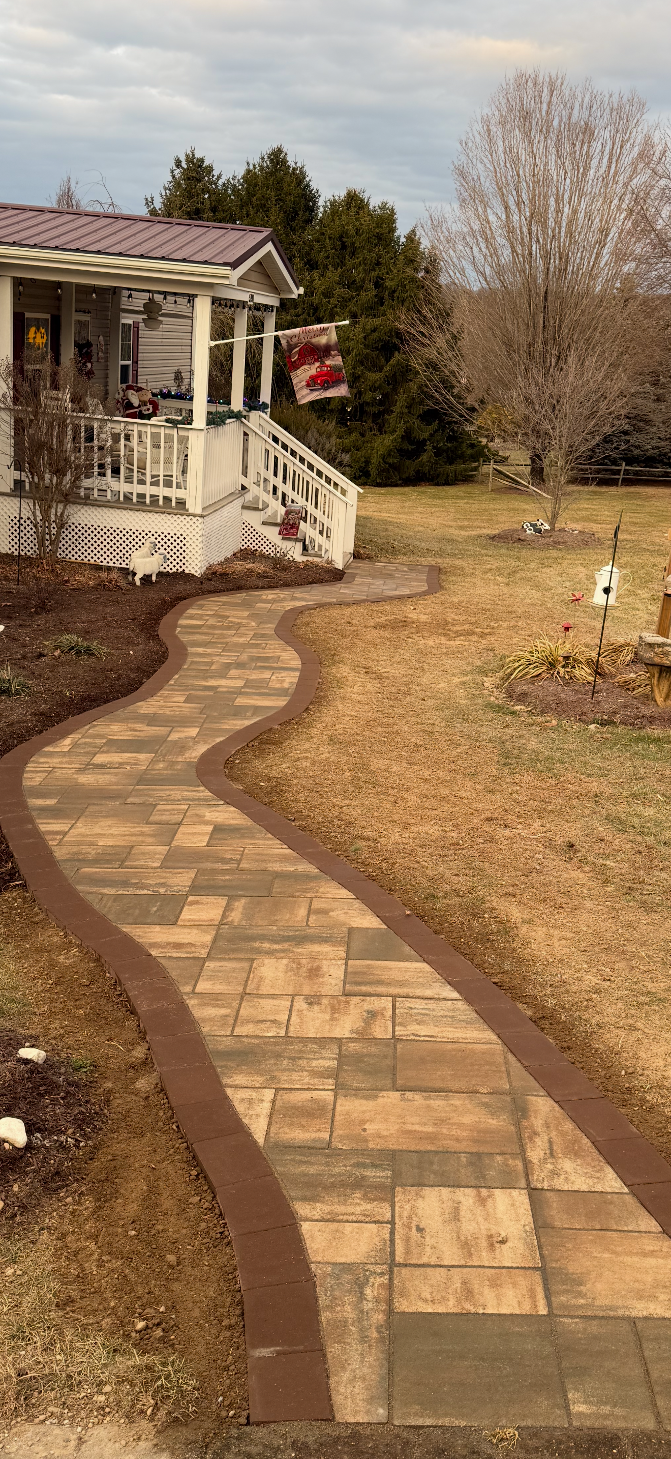 A winding brick pathway in a backyard leads to a porch decorated with holiday ornaments. The yard has some trees, a lawn, and a birdhouse, with a cloudy sky overhead.