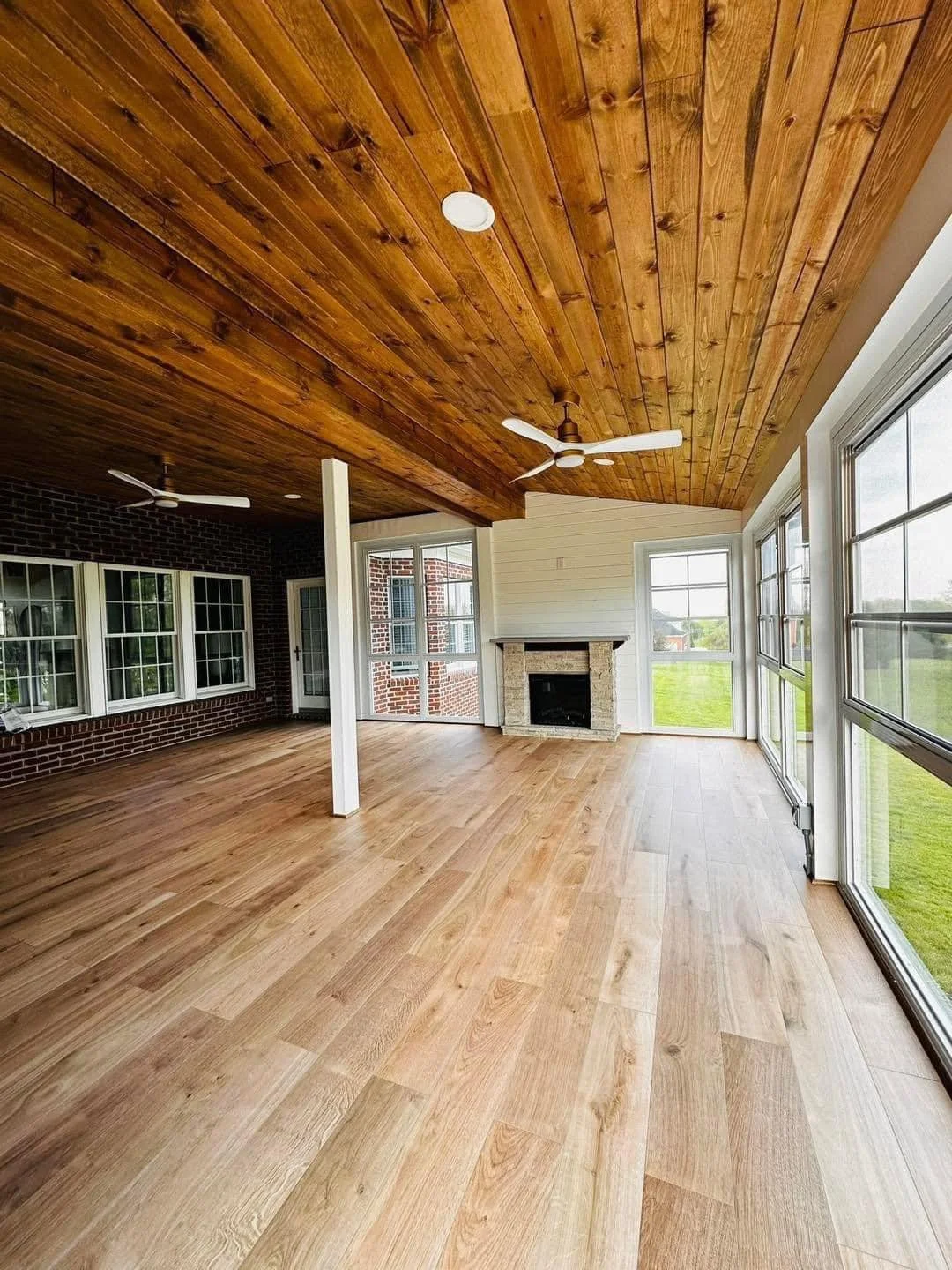 Empty sunroom with light wood flooring, white trim, and large windows, featuring a brick fireplace, wooden ceiling, ceiling fans, and a view of a grassy yard outside.