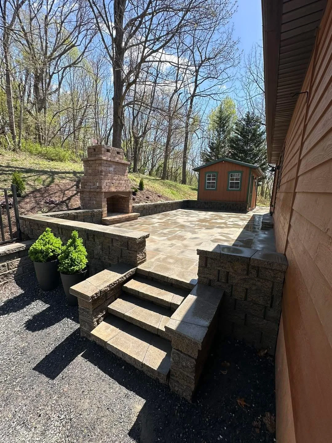A backyard patio with stone steps leading to a paved area, a brick outdoor fireplace, and a small wooden shed surrounded by tall, leafless trees on a sunny day.