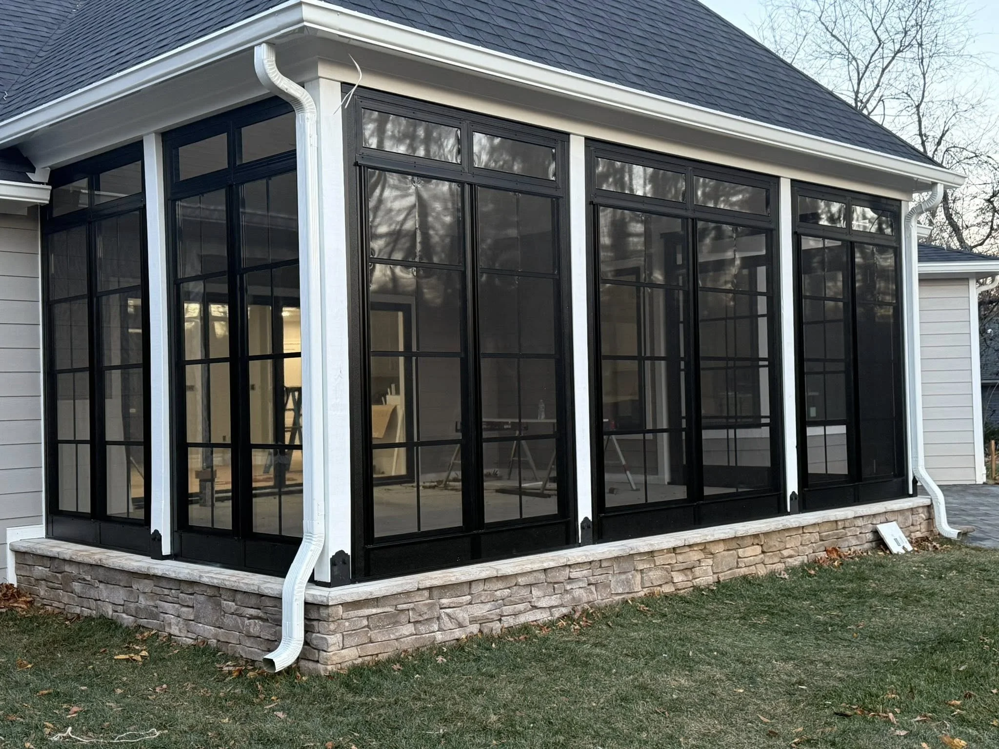 A screened-in porch with black-framed mesh panels on a house with gray siding and stone foundation, showing a view of the interior with some furniture and ladders.