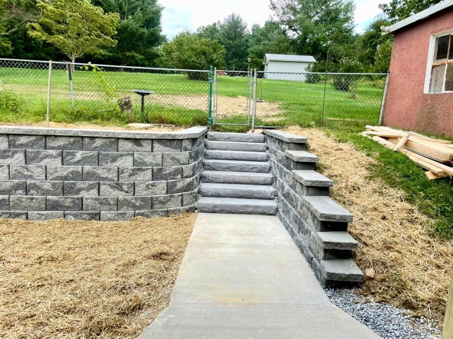 New stone retaining wall and stairs leading up to a fenced backyard with green grass and trees, next to a small pink house.