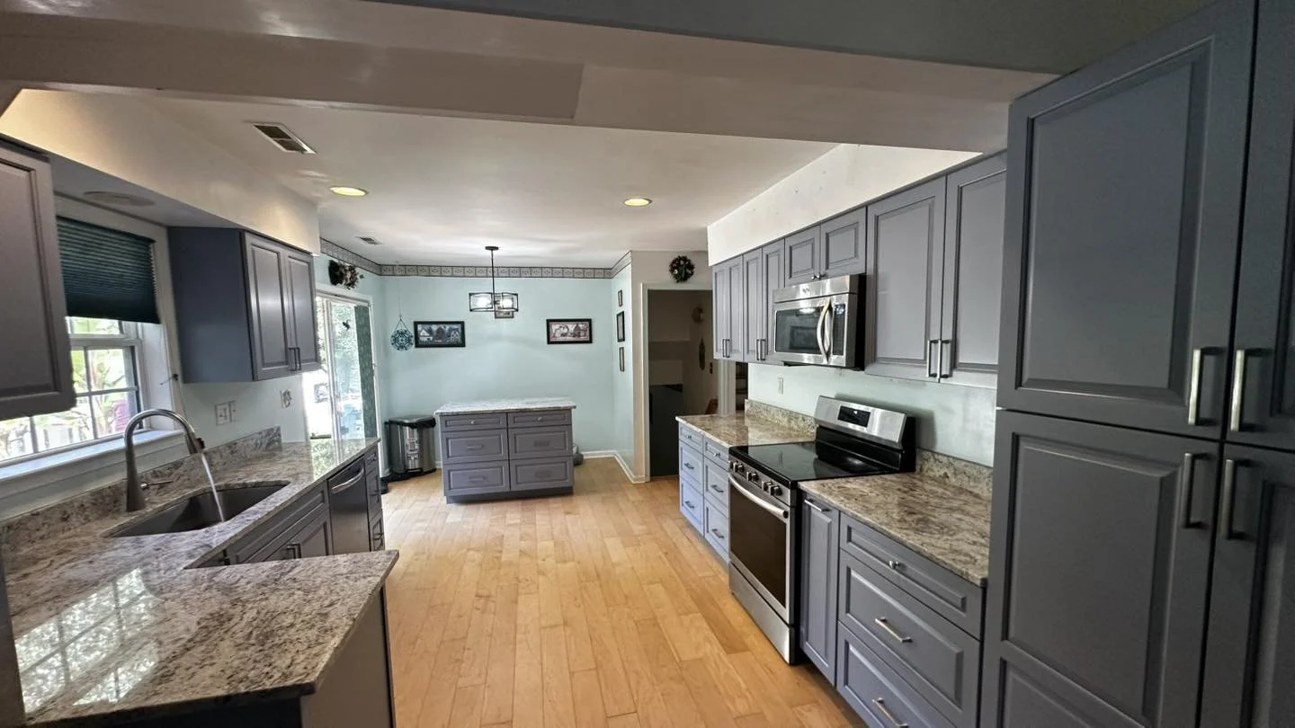 Modern kitchen with gray cabinets, granite countertops, stainless steel appliances, and wooden flooring, with windows and decorative wall art in the background.