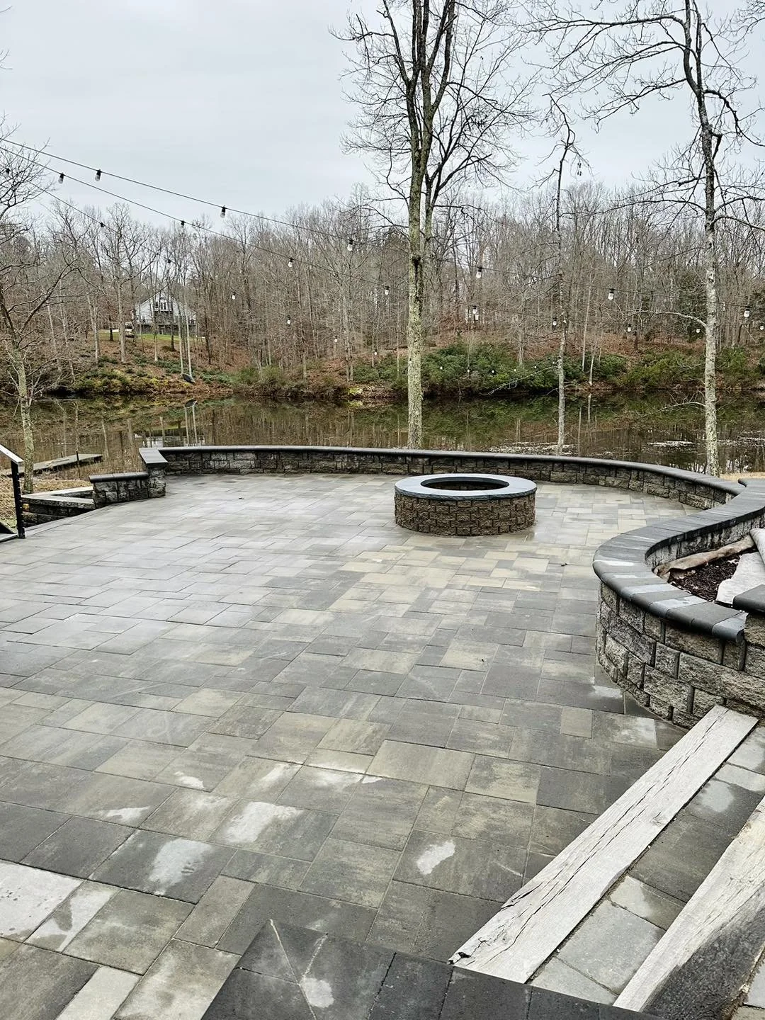 A spacious outdoor patio with stone pavers, a central fire pit, stone retaining walls, overlooking a calm lake with leafless trees and some houses in the background.