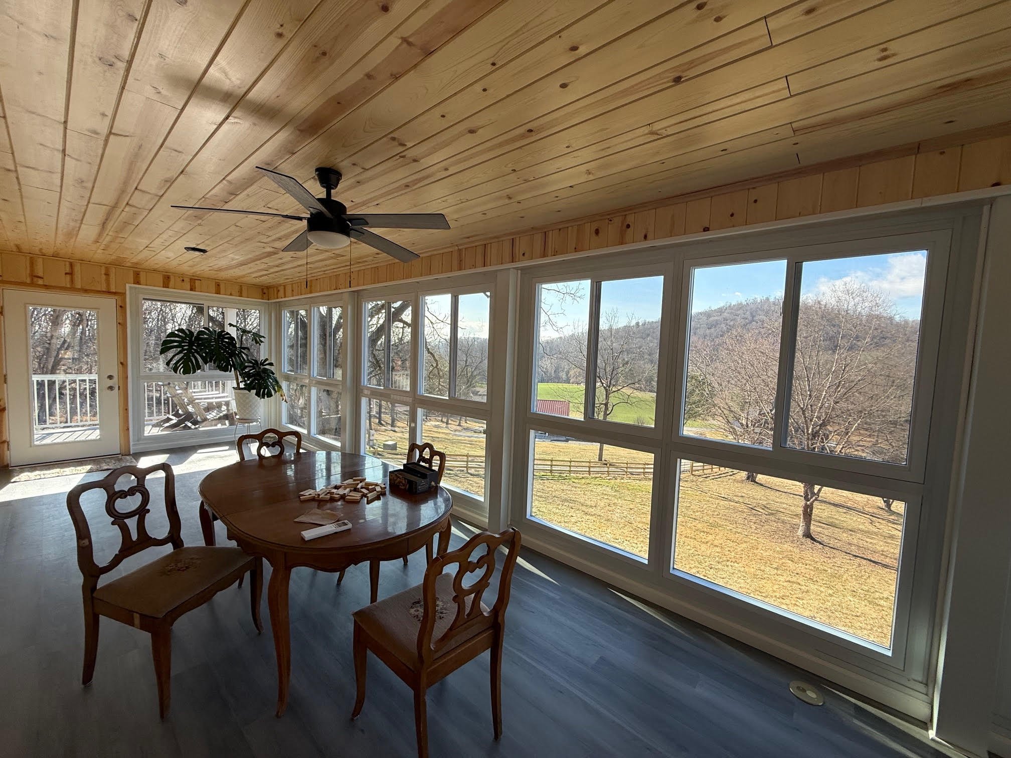 Sunroom with wooden ceiling, large windows, wooden dining table, four chairs, and a ceiling fan, overlooking a yard with trees, fences, and a distant mountain view.