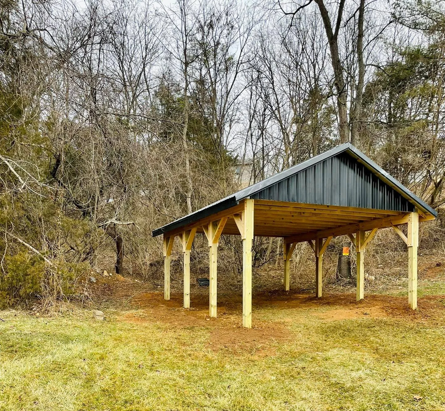 A wooden carport with a metal roof on a grassy yard surrounded by leafless trees in early spring.